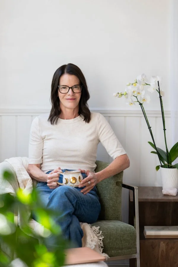 A woman with dark hair, glasses, and a light-colored shirt, sitting on a green chair, holding a mug with gold accents, next to a white orchid plant in a white pot.