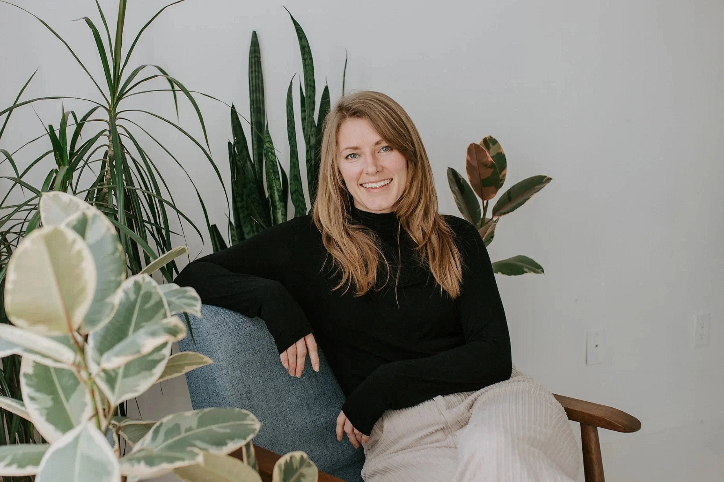 A smiling woman with long, light brown hair, wearing a black top, sitting on a gray couch with a blue cushion, surrounded by green houseplants indoors.