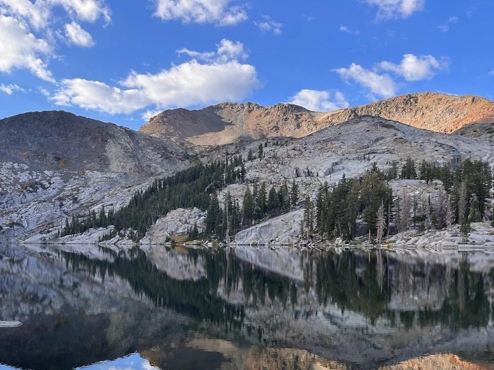 Mountain landscape with rocky peaks, evergreen trees, and a calm lake reflecting the scenery and blue sky with scattered clouds.