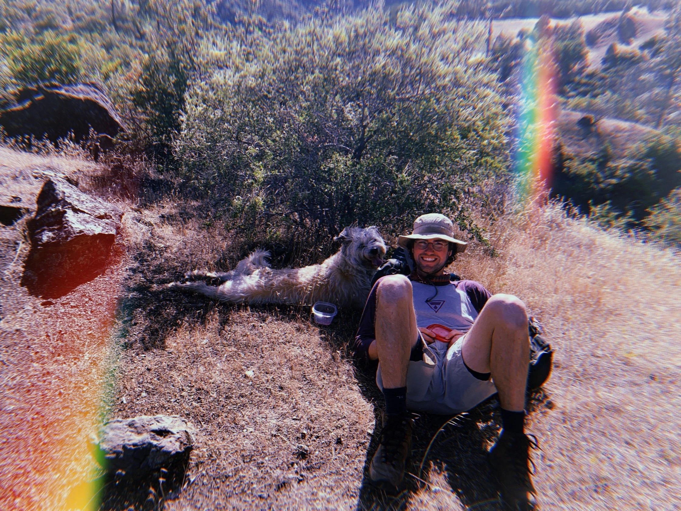 A person and a dog resting on a rocky and dry outdoor trail with bushes and rocks in the background, and a rainbow light streak across the image.