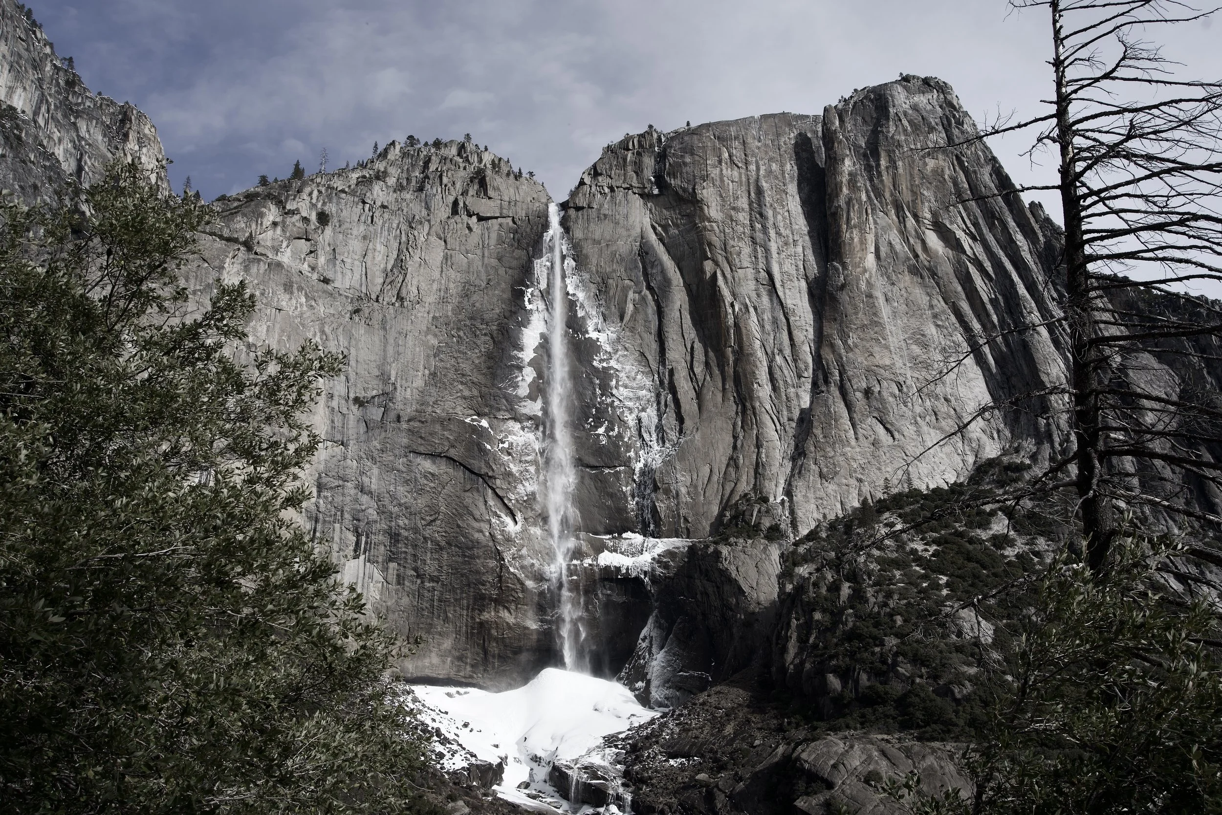 A tall waterfall flowing down a large granite mountain with snow at the bottom, surrounded by tall trees and greenery.