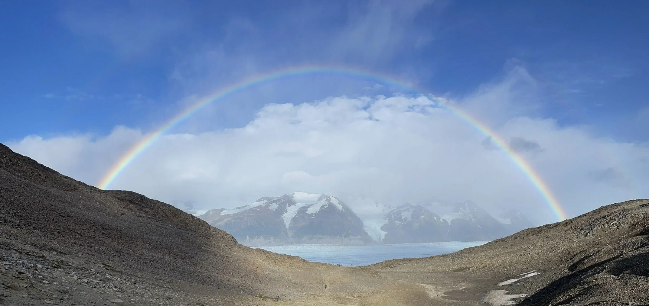 A rainbow over snow-capped mountains, with a clear blue sky and some clouds.