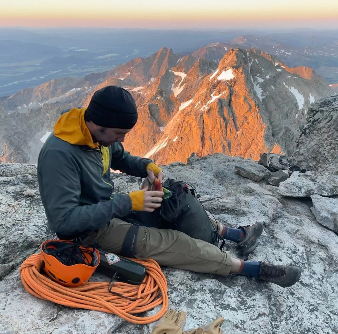 A person sitting on a mountain peak at sunrise, preparing climbing gear with rugged mountain peaks in the background.