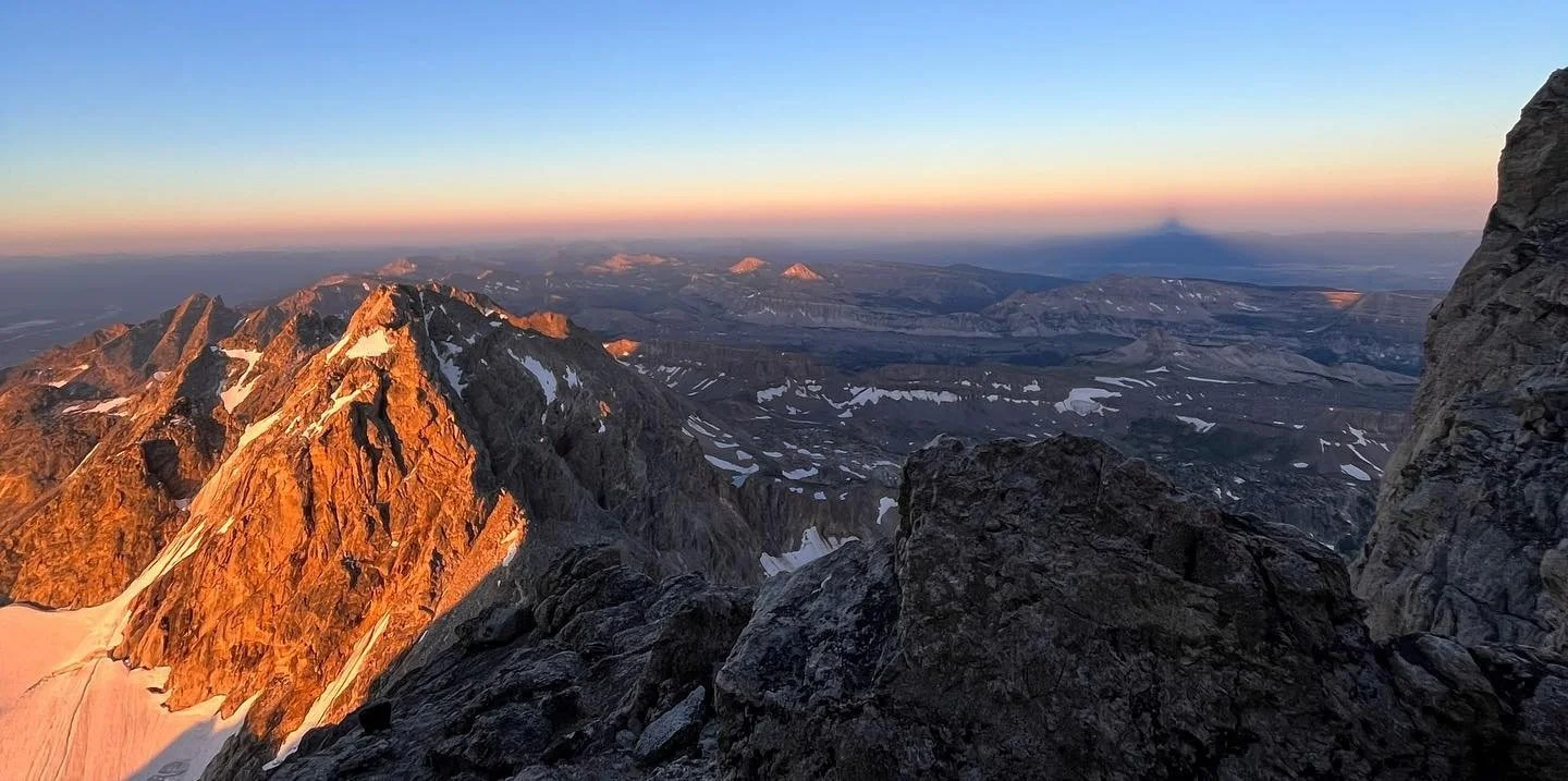Sunlit mountain peaks at sunset with snow patches, viewed from a rocky ridge.