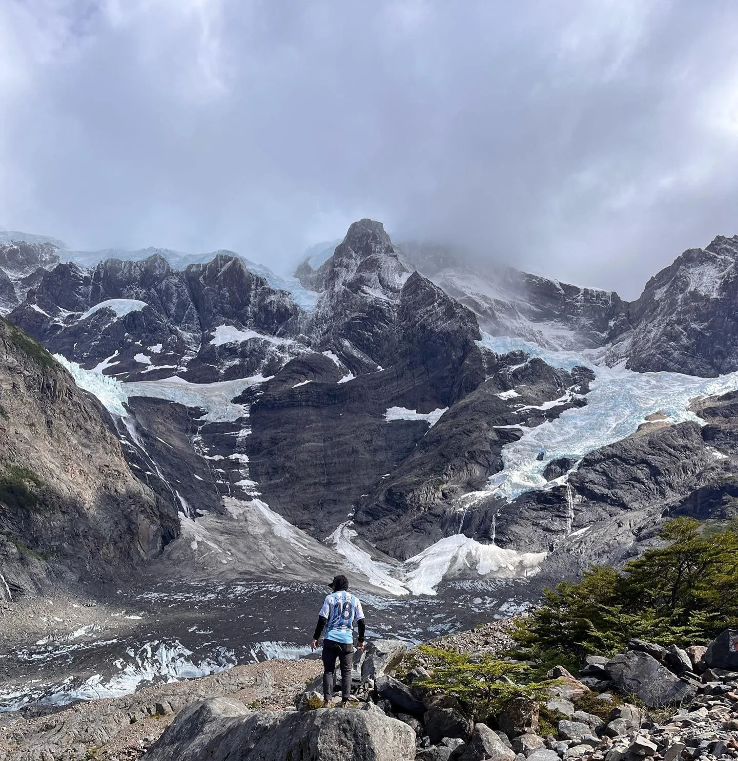 Person standing on rocky terrain looking at snow-capped mountain with glaciers, surrounded by trees and clouds.