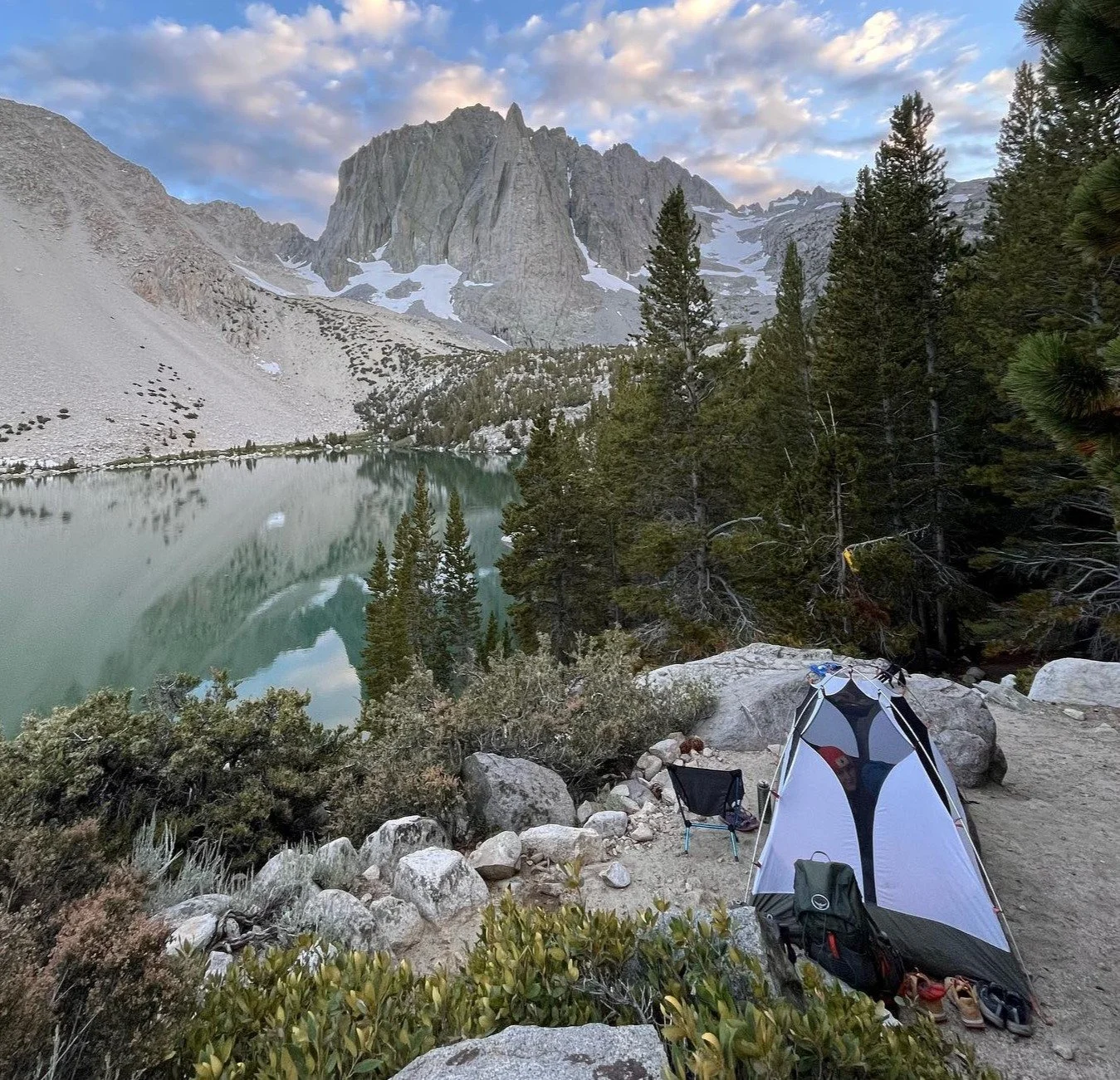 A tent set up on rocky ground next to a forested area by a mountain lake, with tall mountains and partly cloudy sky in the background.