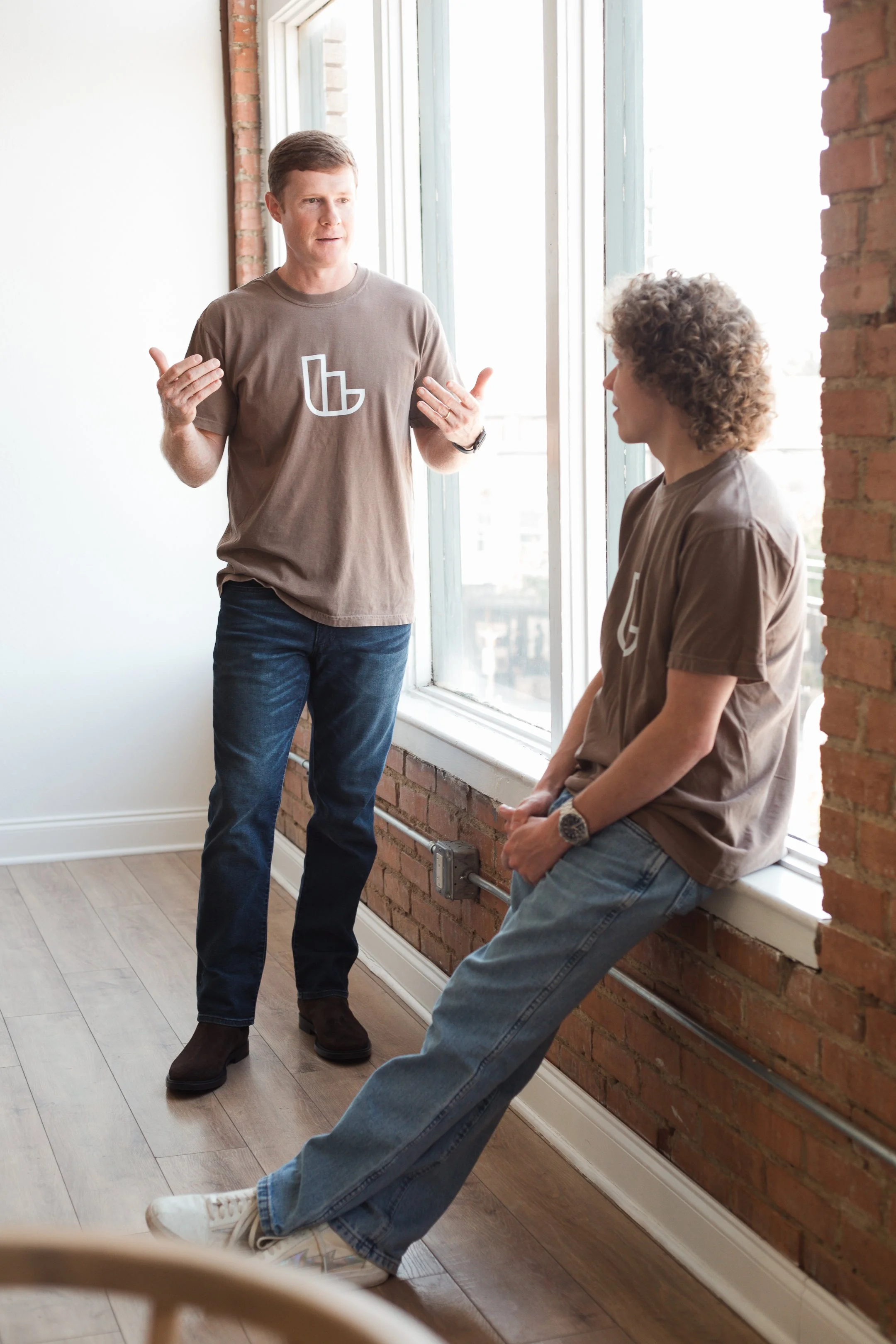 Two men having a conversation in a bright room with a large window and brick wall. One man is standing and gesturing with his hands, while the other is sitting on the window sill, listening.