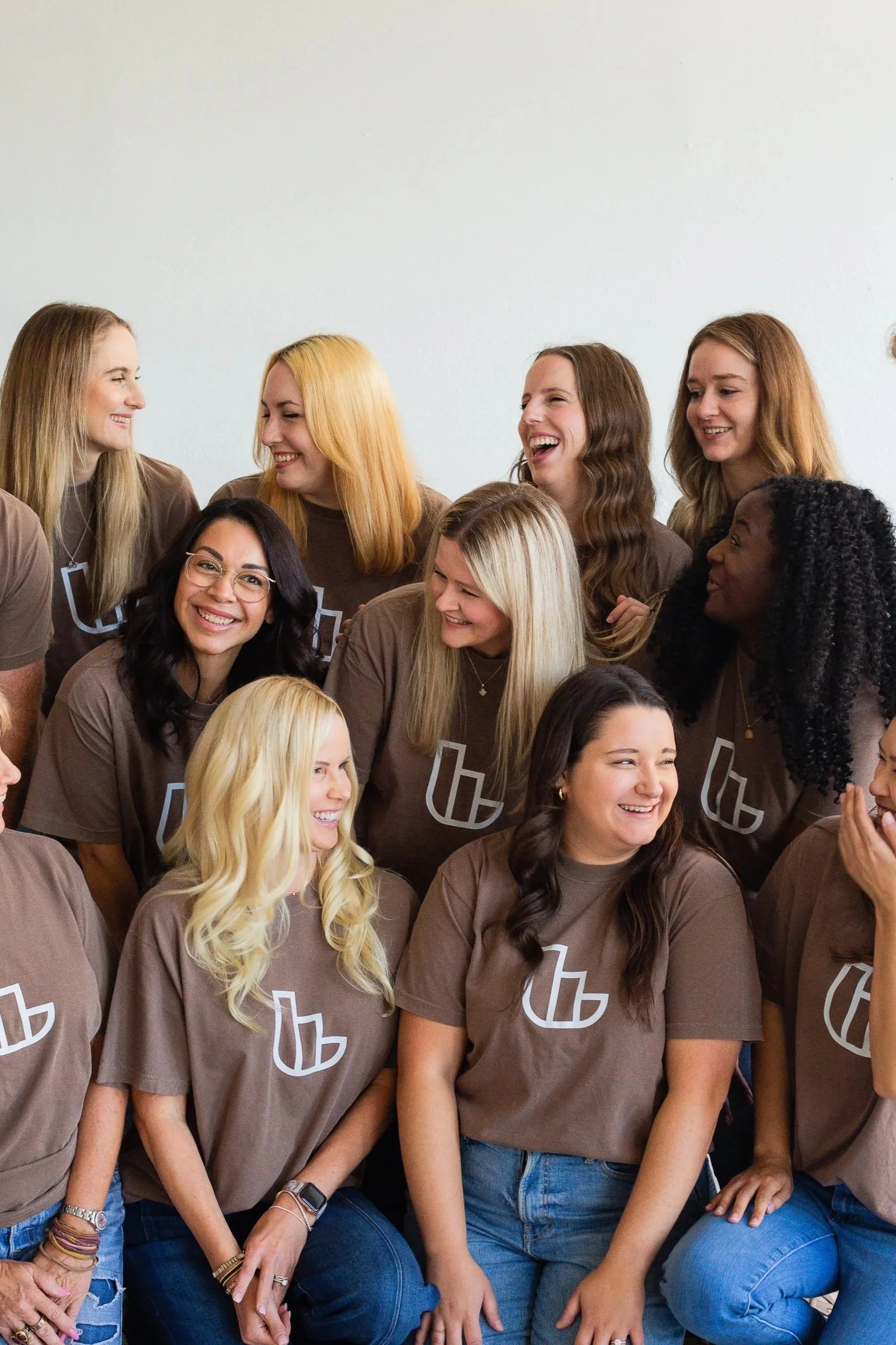 Group of women wearing matching brown T-shirts with a white logo, smiling and laughing together indoors.
