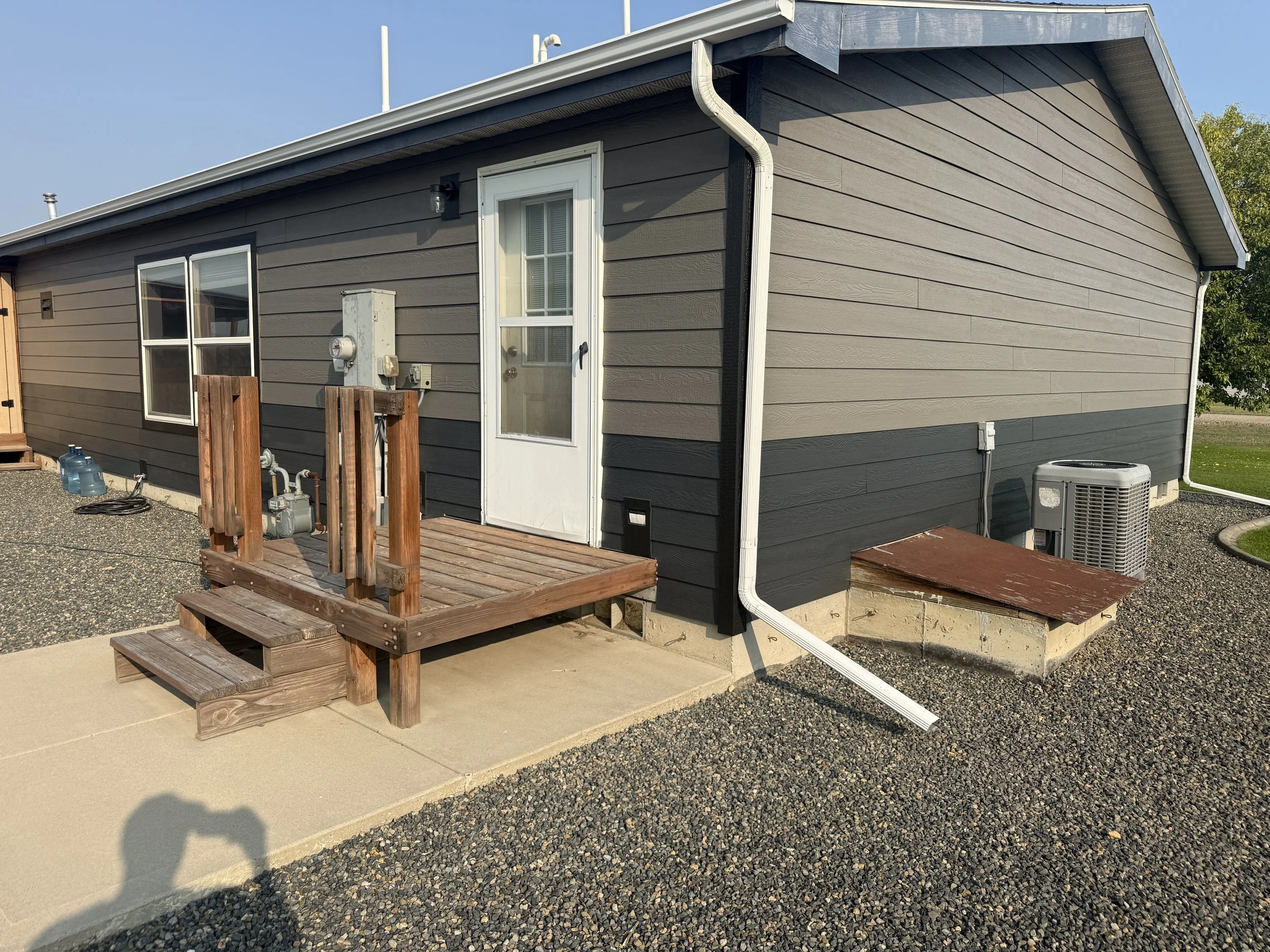 Backyard of a house with a small wooden deck, white door, gray and black siding, and gravel yard. There are two small platforms on the ground, an air conditioning unit, and a gray shed.