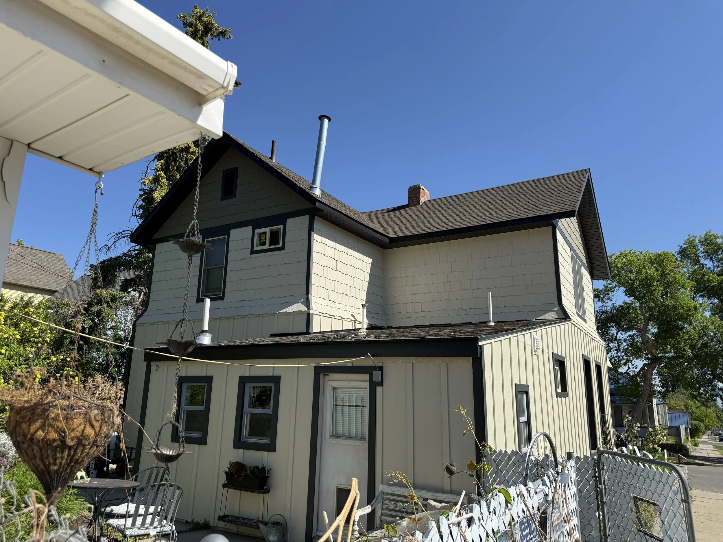 A two-story house with beige siding and dark trim under a clear blue sky, seen from the backyard with a fence and garden.