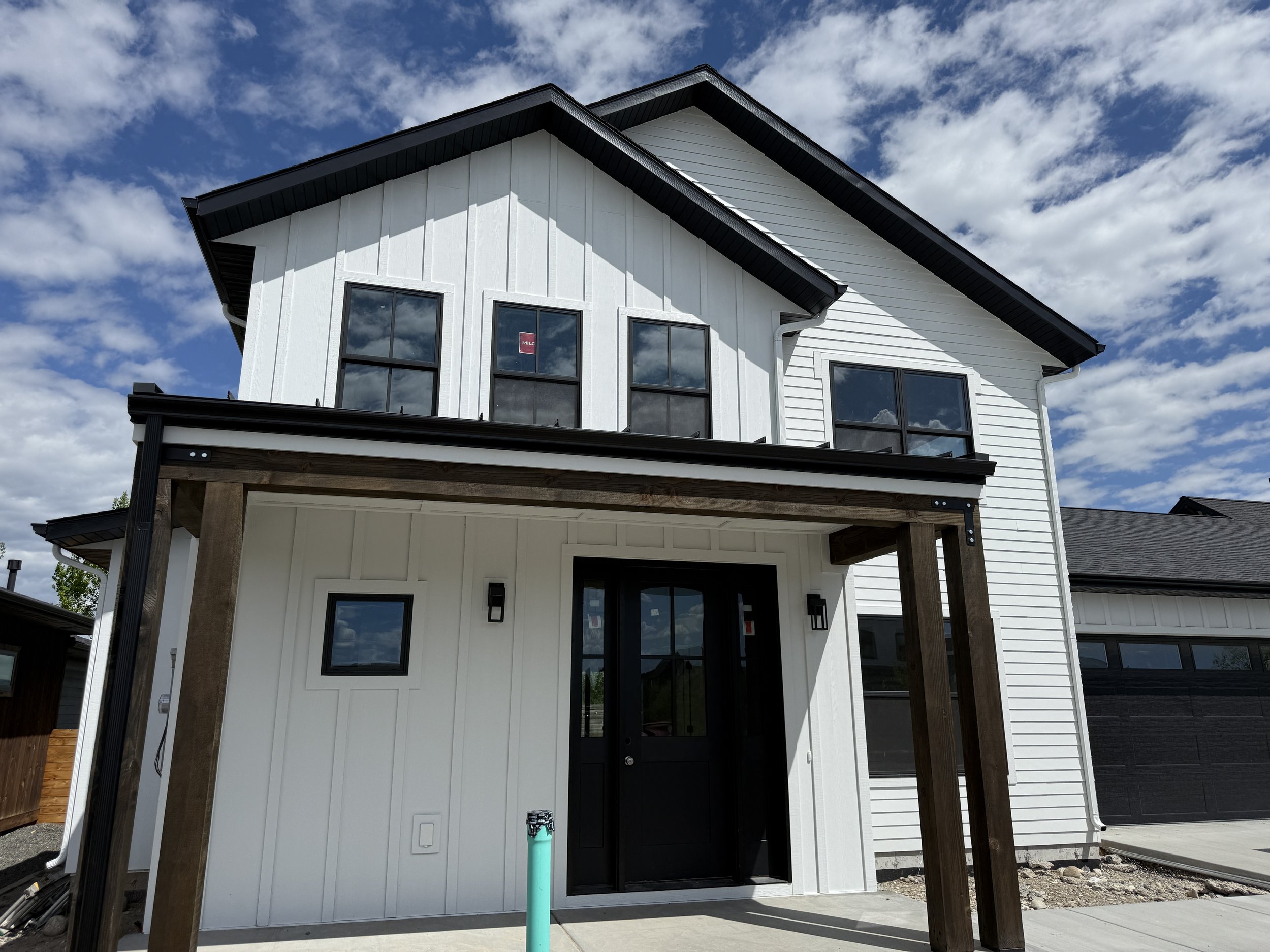 New two-story white house with black trim and black front door, under construction, with exposed wooden beams support over front entrance, and a new PVC pipe in front.
