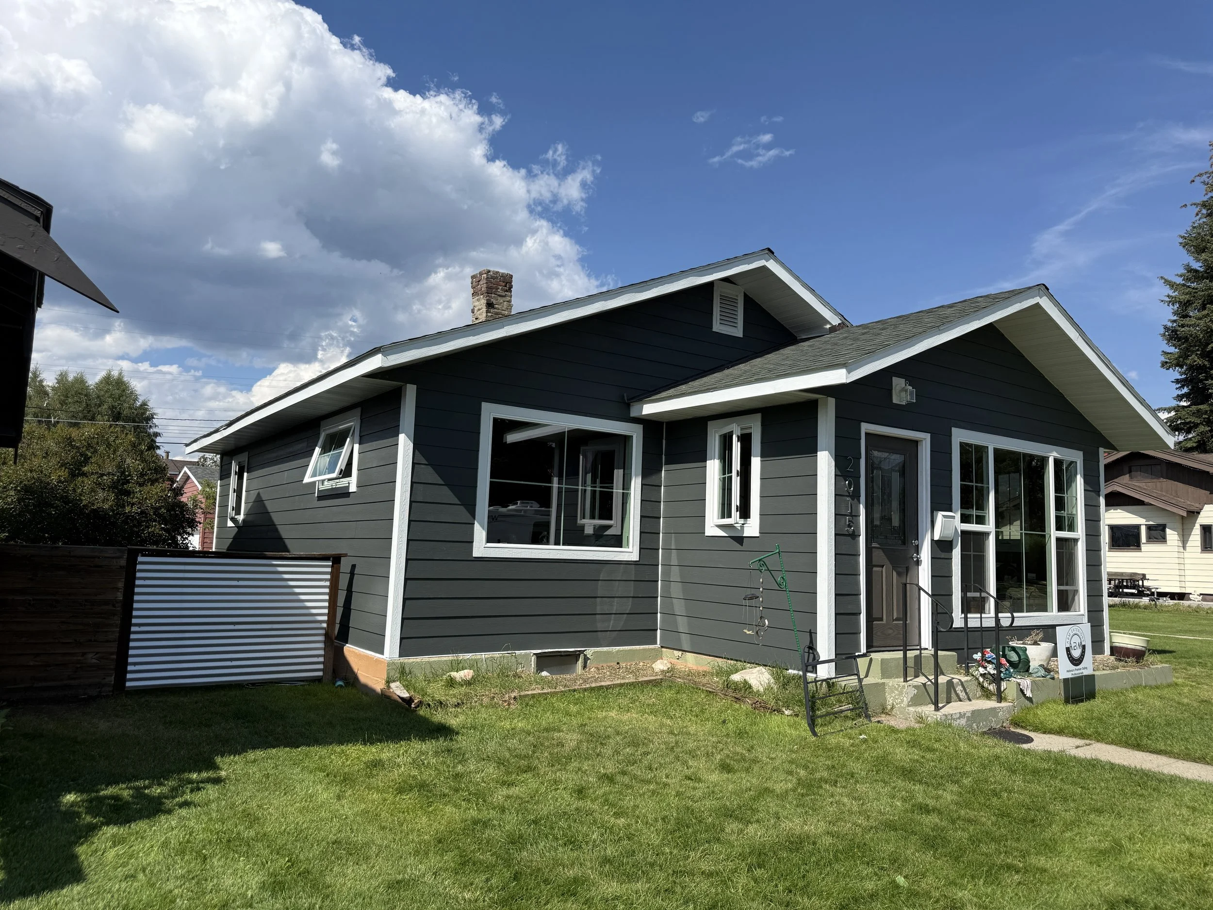 A modern, dark gray house with white trim and a small front yard under a partly cloudy sky.