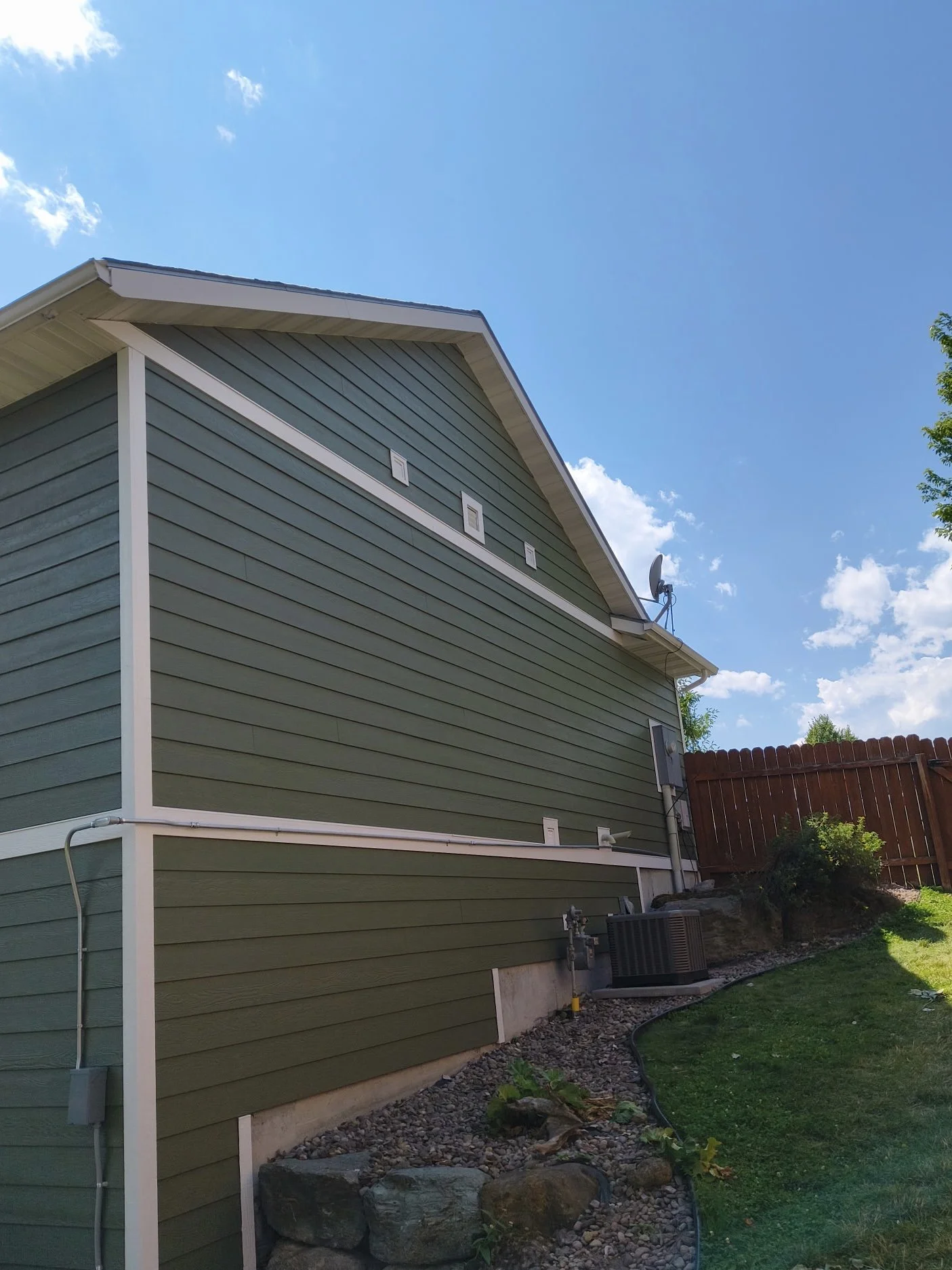 Side view of a green house with white trim, a satellite dish on the roof, and a wooden fence in the yard under a partly cloudy sky.
