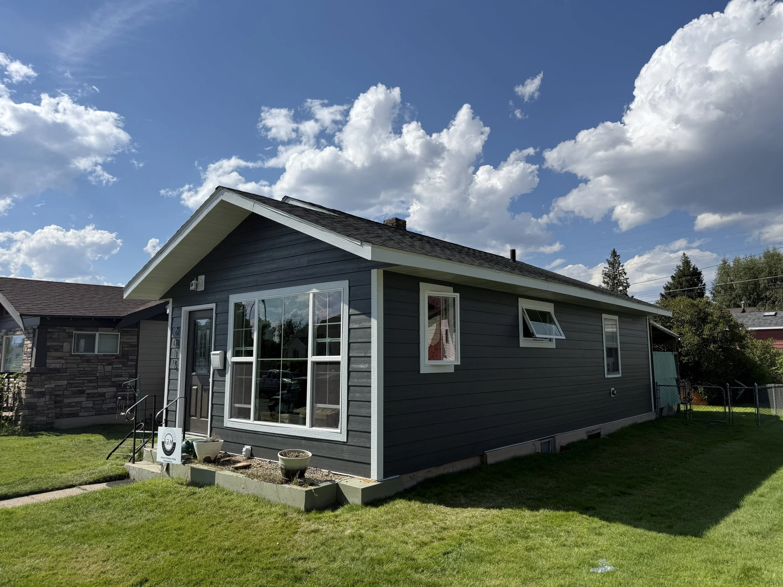 Front view of a dark gray house with large windows, a small porch with steps, and a well-maintained lawn under a partly cloudy sky.