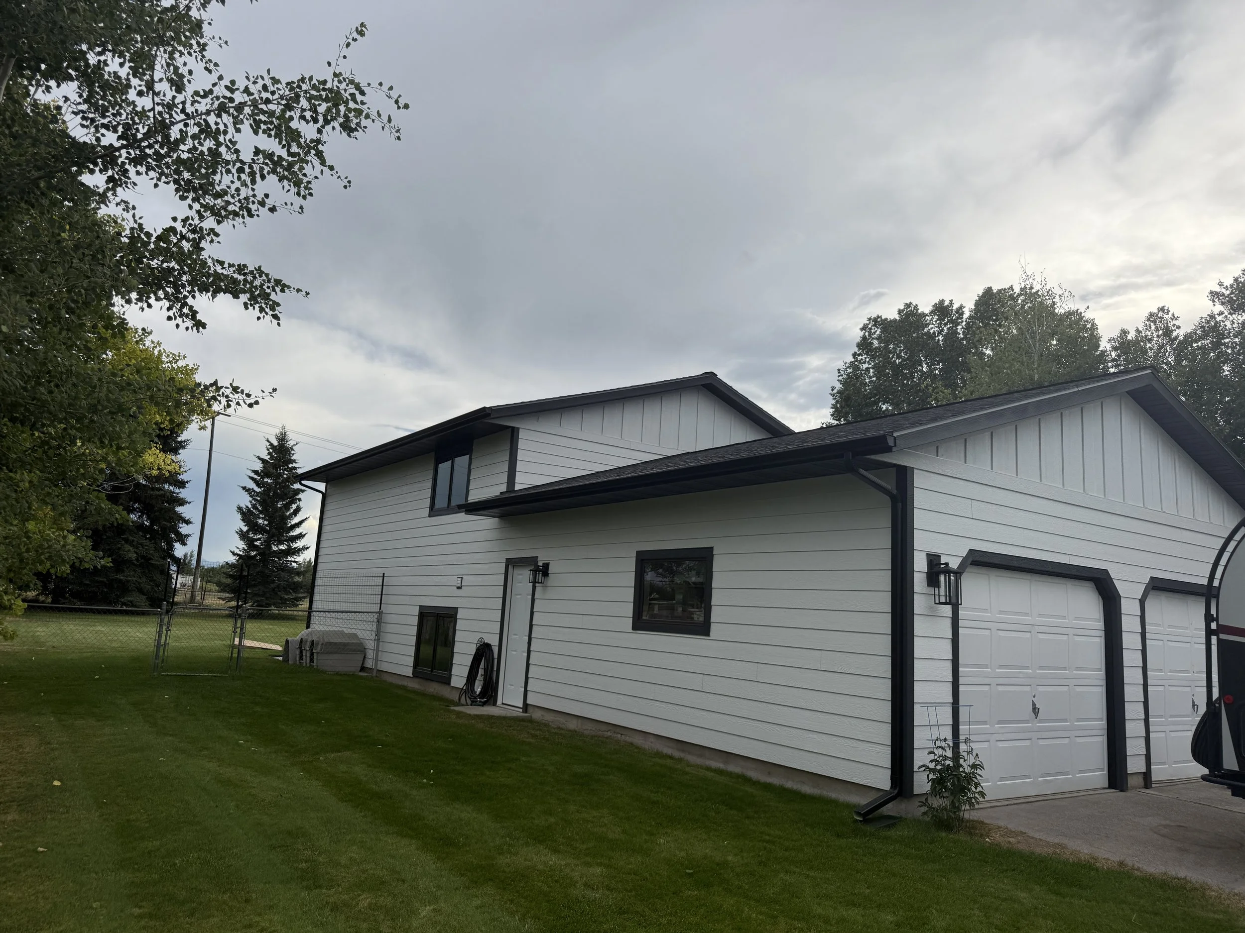 A white two-story house with black trim and garage doors, with a green lawn and trees in the background under a partly cloudy sky.