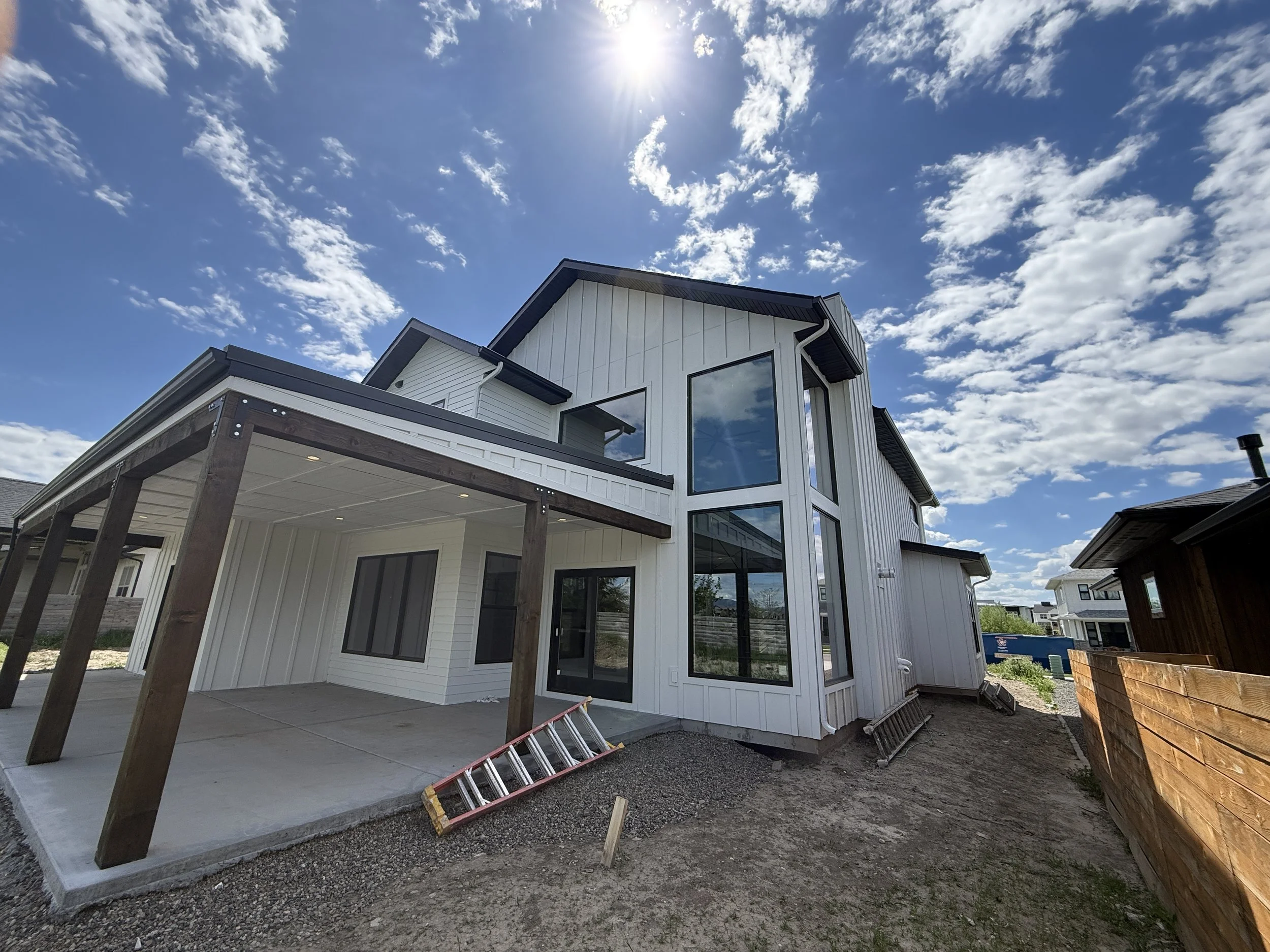 New modern two-story house with white siding, large windows, and a covered outdoor patio under construction, on a sunny day with a partly cloudy sky.