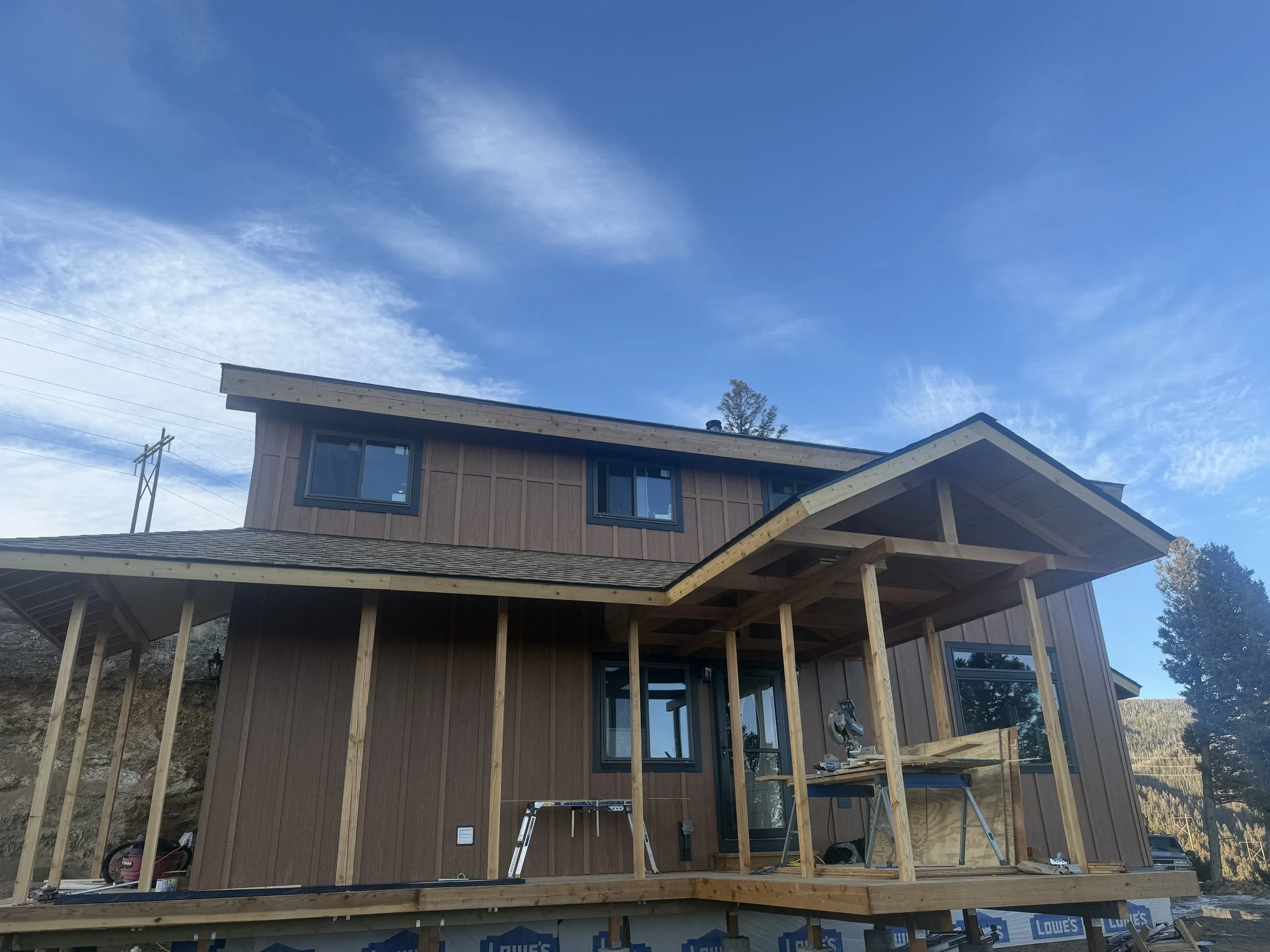 A two-story house under construction with wooden framing and a partially completed covered porch, set against a blue sky with some clouds and trees in the background.