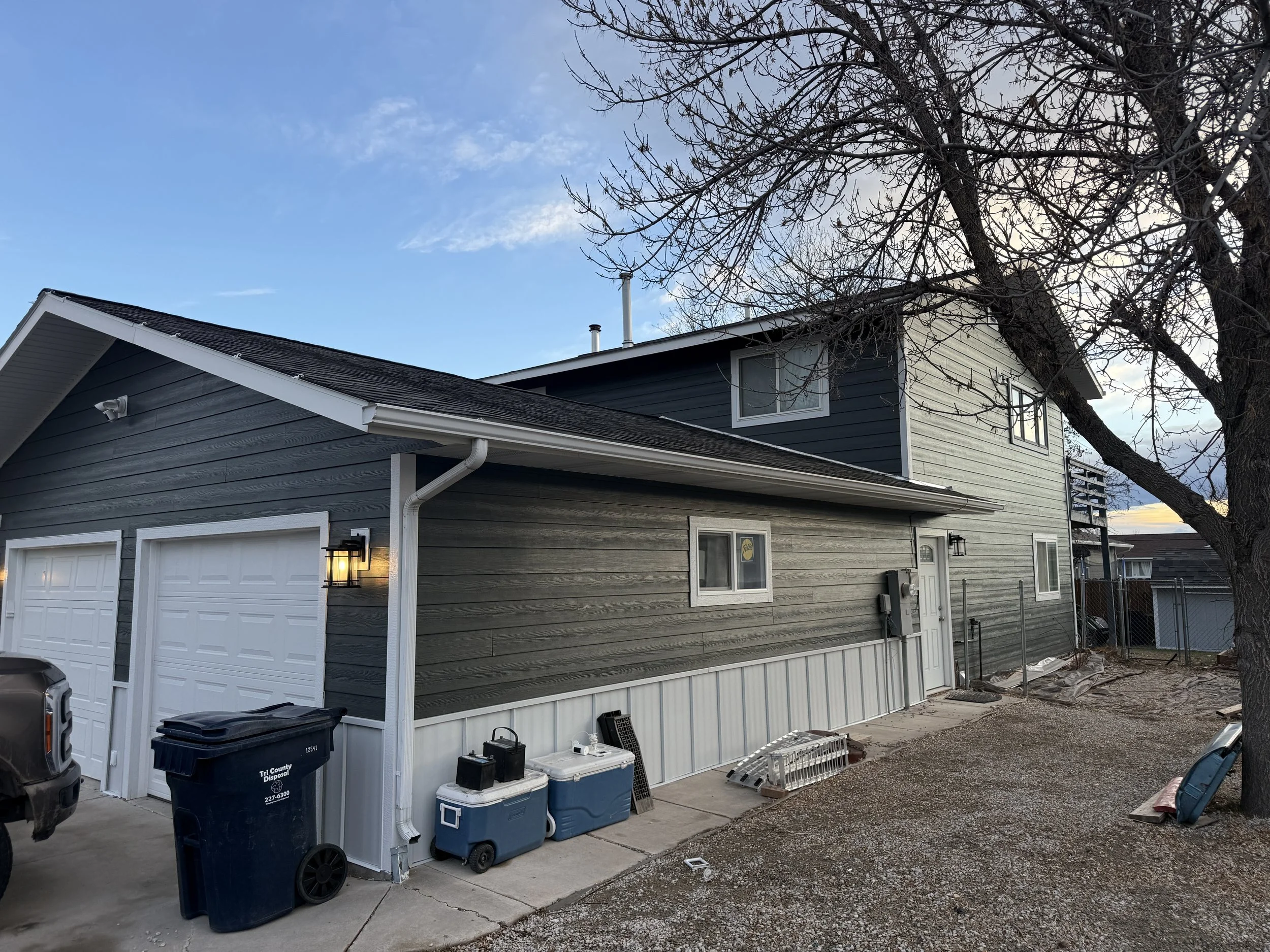 Exterior view of a two-story house with gray and white siding, a garage, and leafless trees, during dusk or early evening.