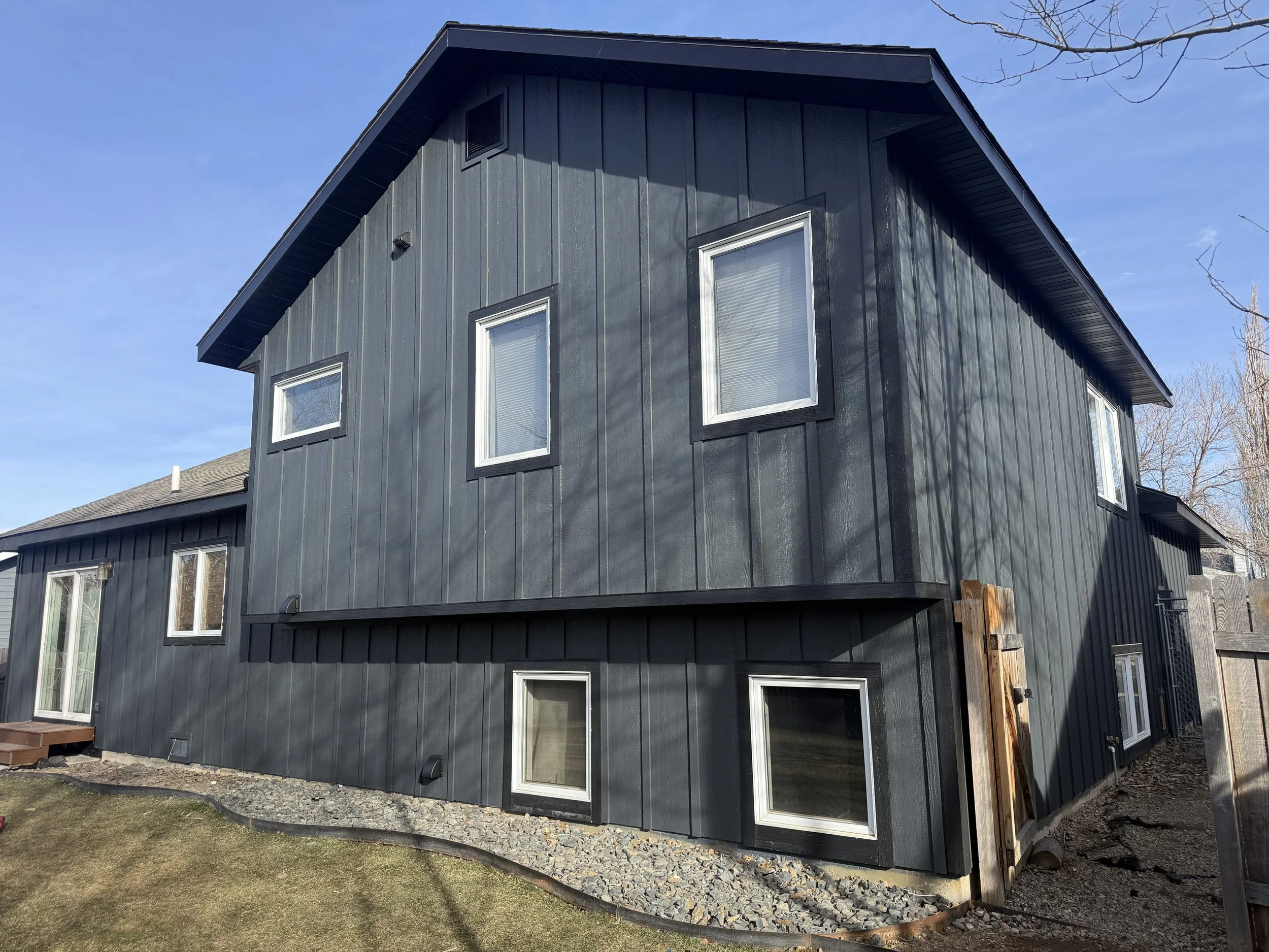 Black two-story house with vertical siding, multiple windows, and a small fenced area at the right side, with a gravel path and a small front yard.