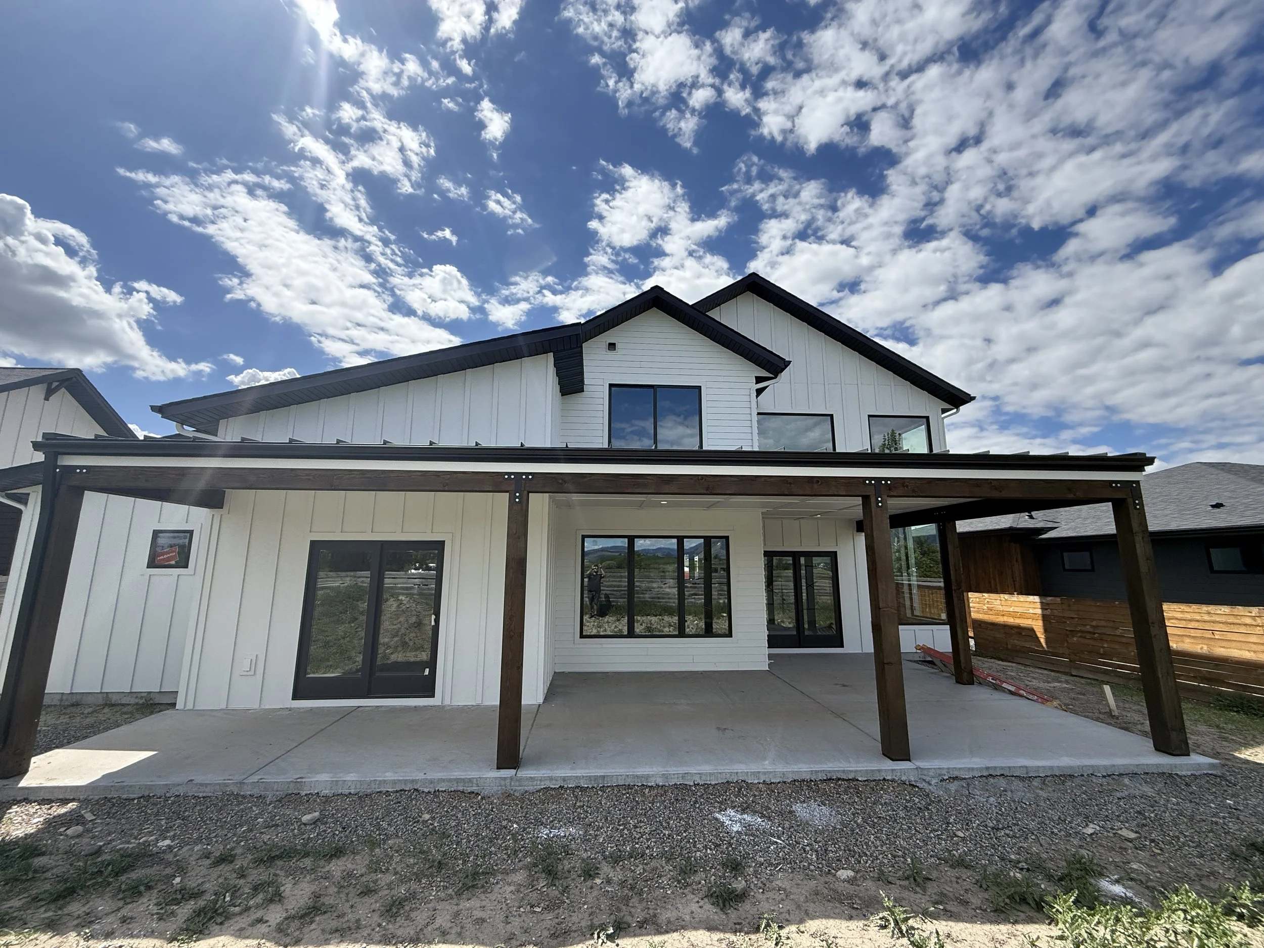 New two-story house with white siding and black trim, featuring a covered patio with wooden beams and large windows under a blue sky with scattered clouds.