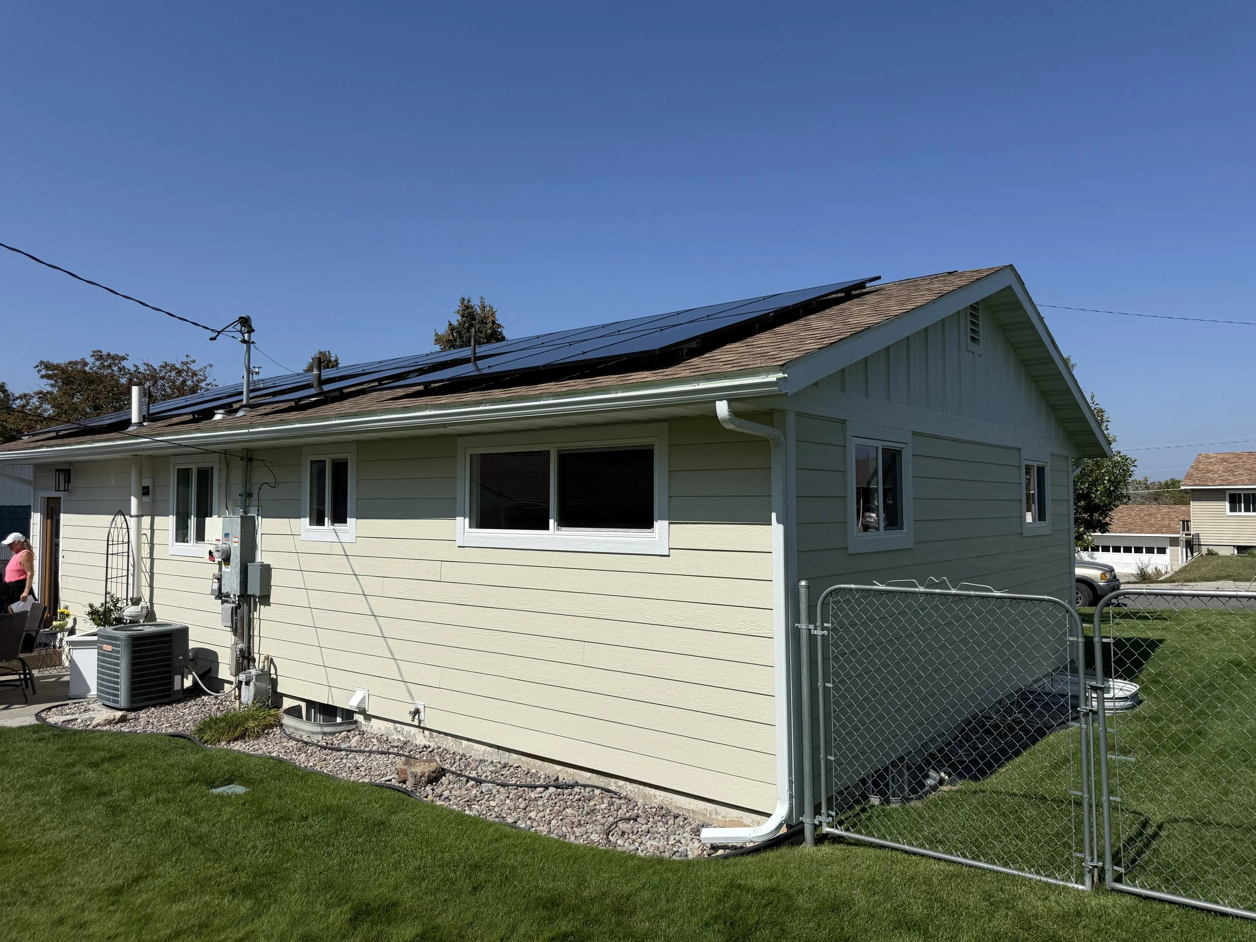 Side view of a beige house with solar panels on the roof, multiple windows, a small backyard with a chain-link fence, and an air conditioning unit.