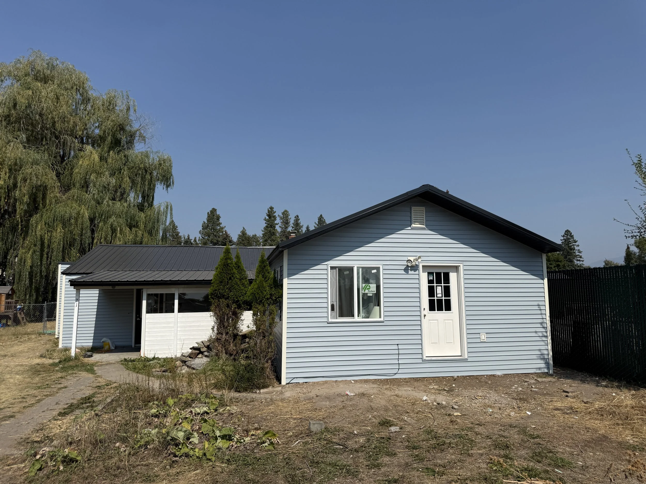A light blue house with white door and window, small front yard with dry grass, and surrounded by trees and a chain-link fence under a clear blue sky.