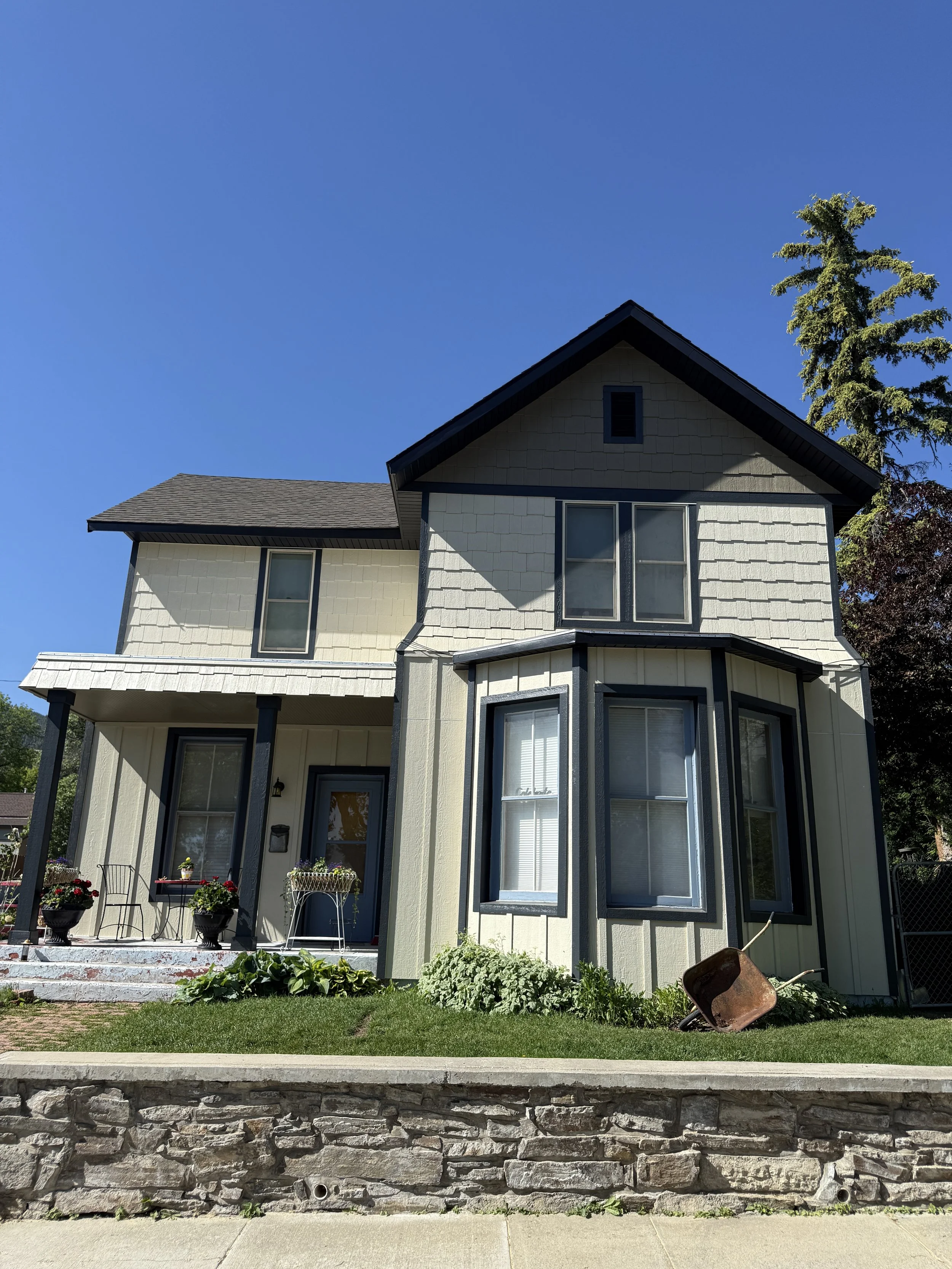 A two-story house with a porch, light-colored exterior walls, dark trim, and a garden in front. There are potted plants on the porch, a wheelbarrow resting on the lawn, and a clear blue sky above.