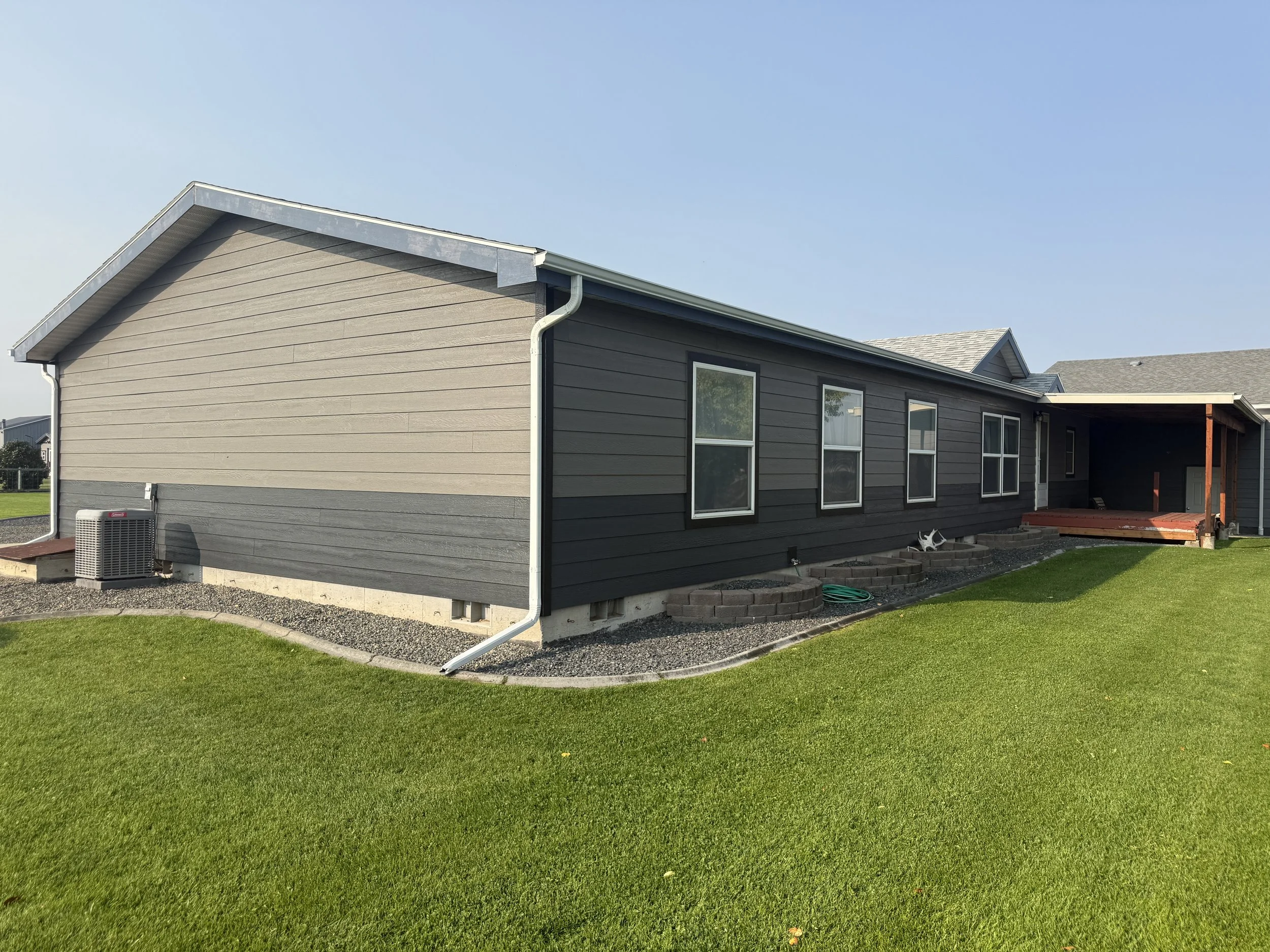 Side view of a house with dark gray siding, several windows, a small porch, green lawn, and clear sky.