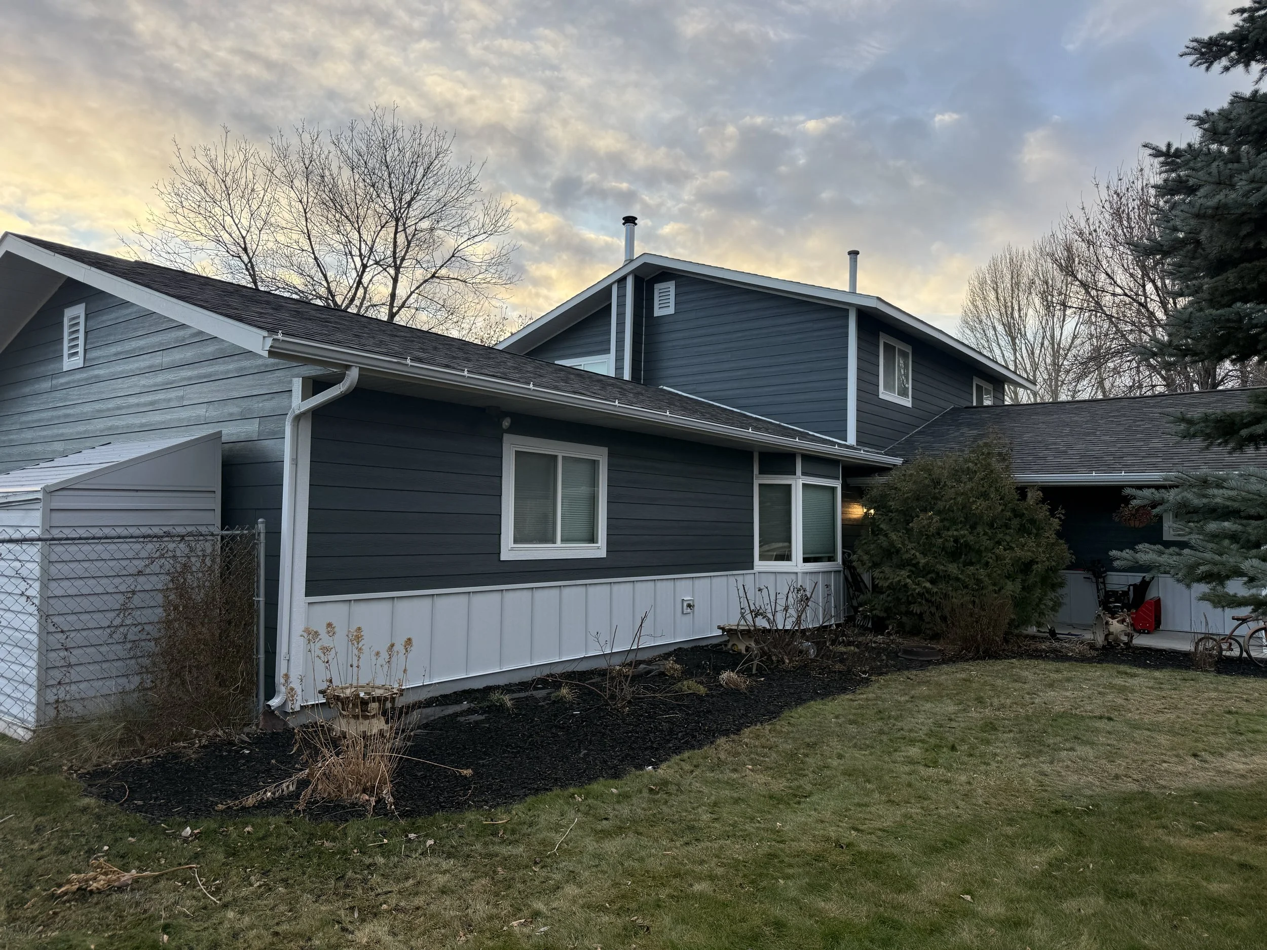 The back of a blue house with white trim, a garden with plants and bushes, a lawn, and trees in the background during sunset.