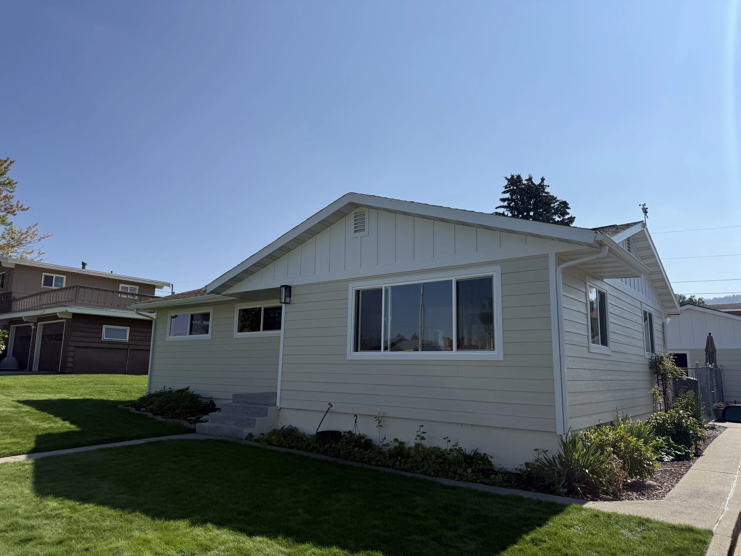 A single-story house with beige siding, a large front window, and concrete steps leading to the front door, situated on a well-manicured lawn under a clear blue sky.