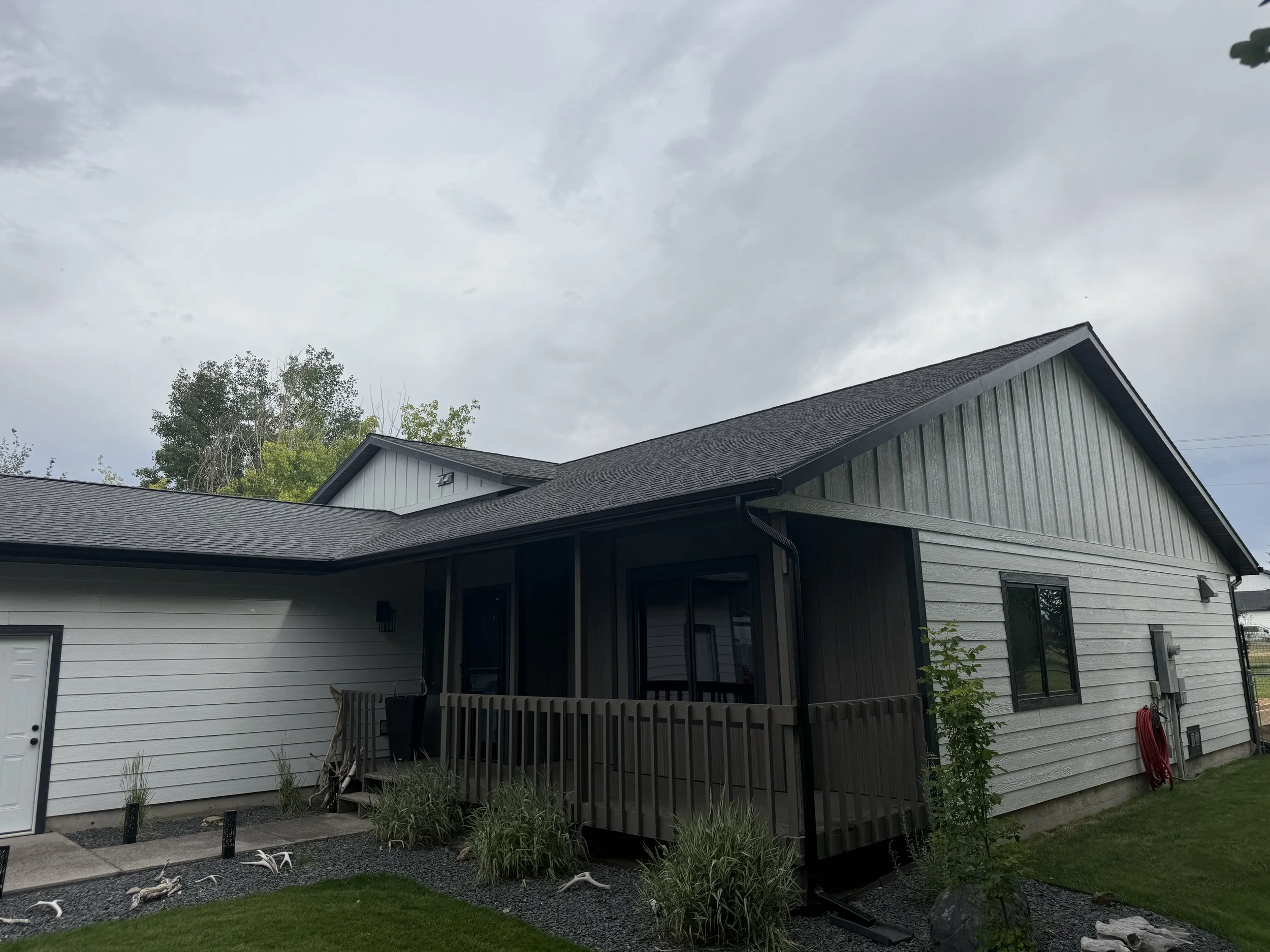 Exterior view of a modern house with gray siding, a porch with a wooden railing, multiple windows, a garden area with plants, and a cloudy sky overhead.