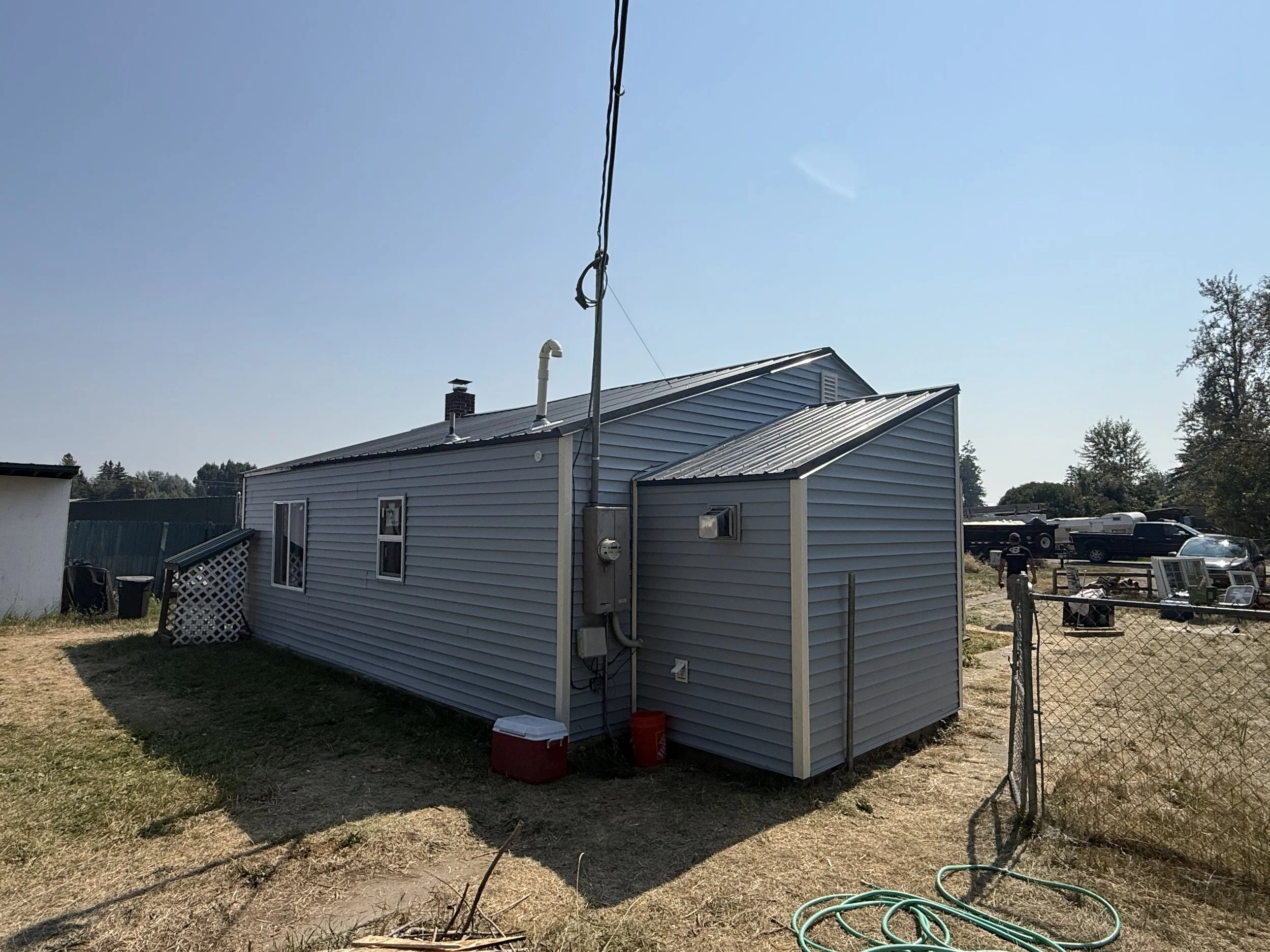 Side view of a light blue suburban house with a metal roof, two small windows, and utility connections on the side. The yard has patches of grass and dirt, a red cooler, a garden hose, and a chain-link fence. In the background, there are parked cars,