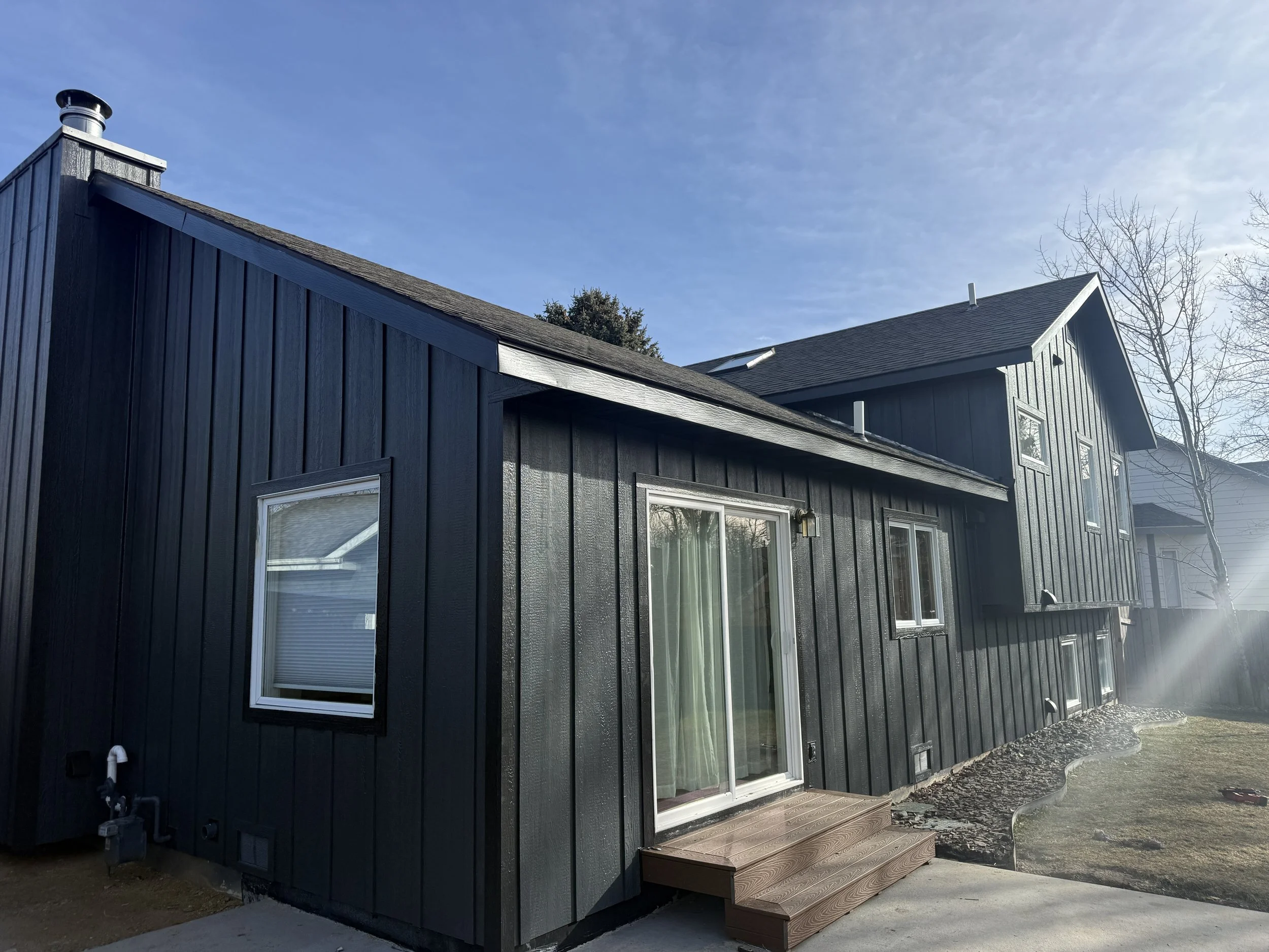 View of a modern black two-story house with vertical siding, patio door, steps, windows, and a backyard with grass and a tree under a blue sky.