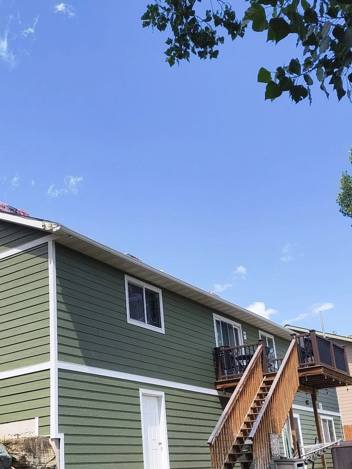 Back view of a two-story house with green siding, white trim, and a wooden staircase leading to a balcony. The sky is clear with a few clouds, and some tree branches are visible at the top.