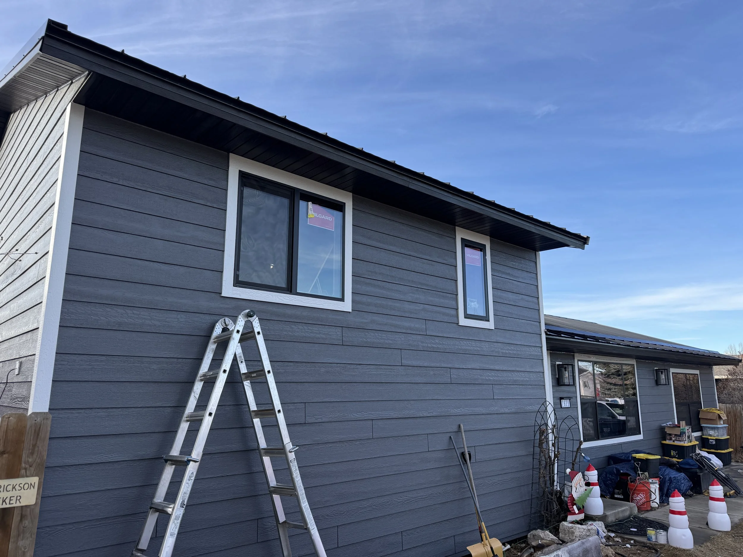Side view of a gray house with white trim, two windows, and a ladder leaning against the wall. The yard has various tools, decorations, and boxes, with Christmas-themed ornaments and traffic cones outside.