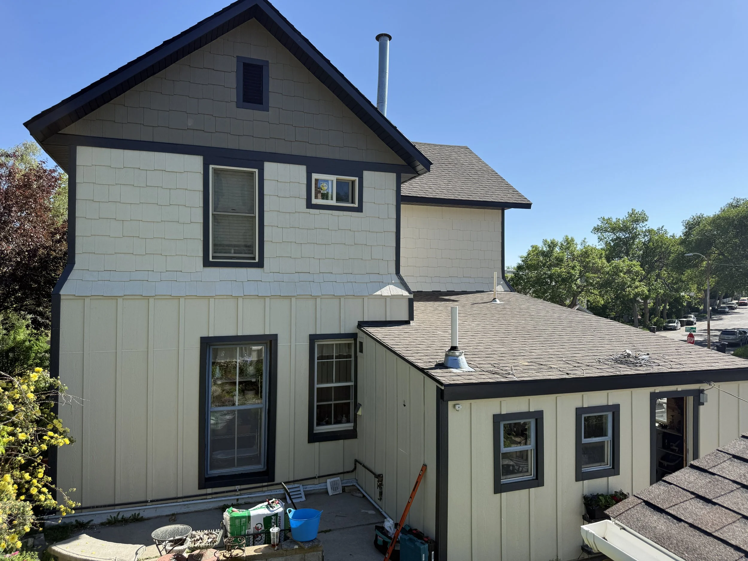 A multi-story house with beige and dark blue exterior siding, with several windows, some with blinds, and a sloped roof with visible vents and chimney pipes. The house is surrounded by trees and a clear blue sky.
