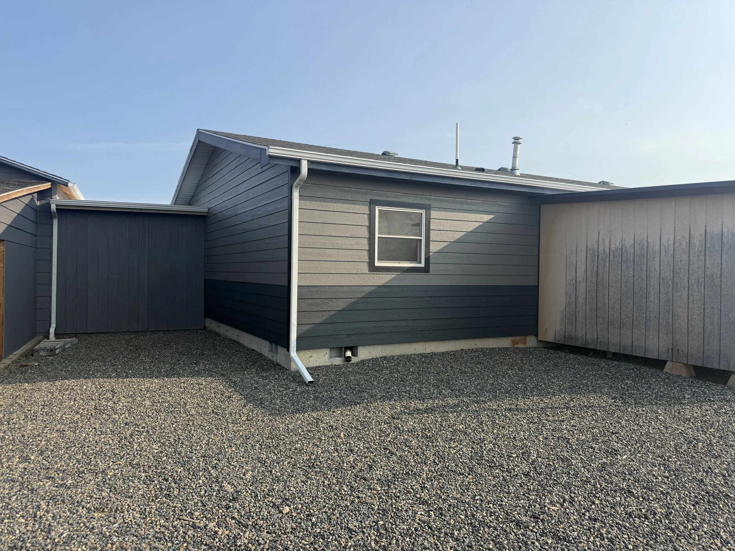 Exterior view of a house with gray siding, gravel ground, and a clear blue sky.
