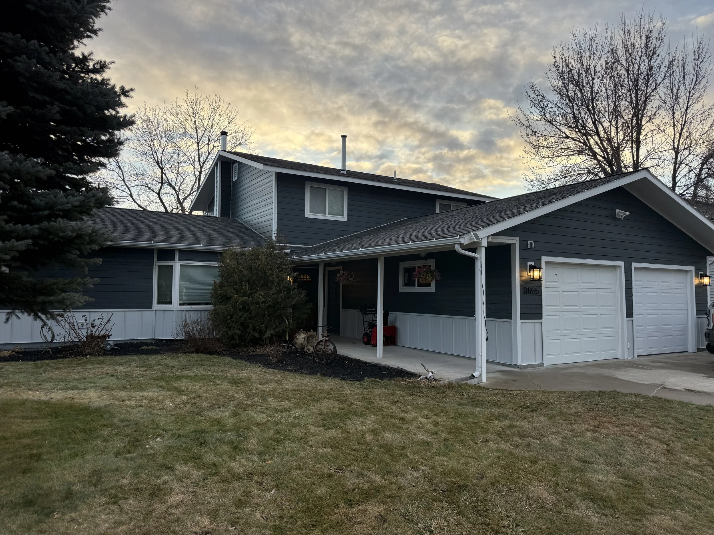 A two-story blue house with white trim and a double garage, in a suburban neighborhood during dusk with a partly cloudy sky.