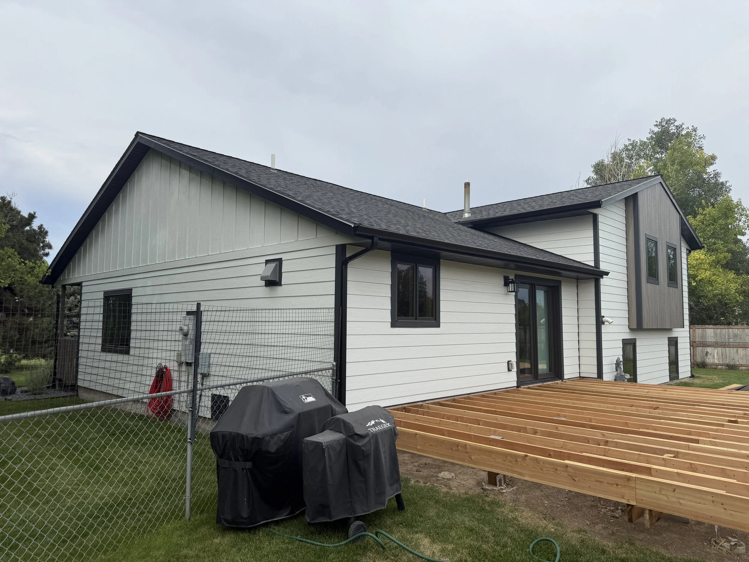 Backyard of a house under construction with a wooden deck frame, grill covered with a black cover, and chain-link fence.