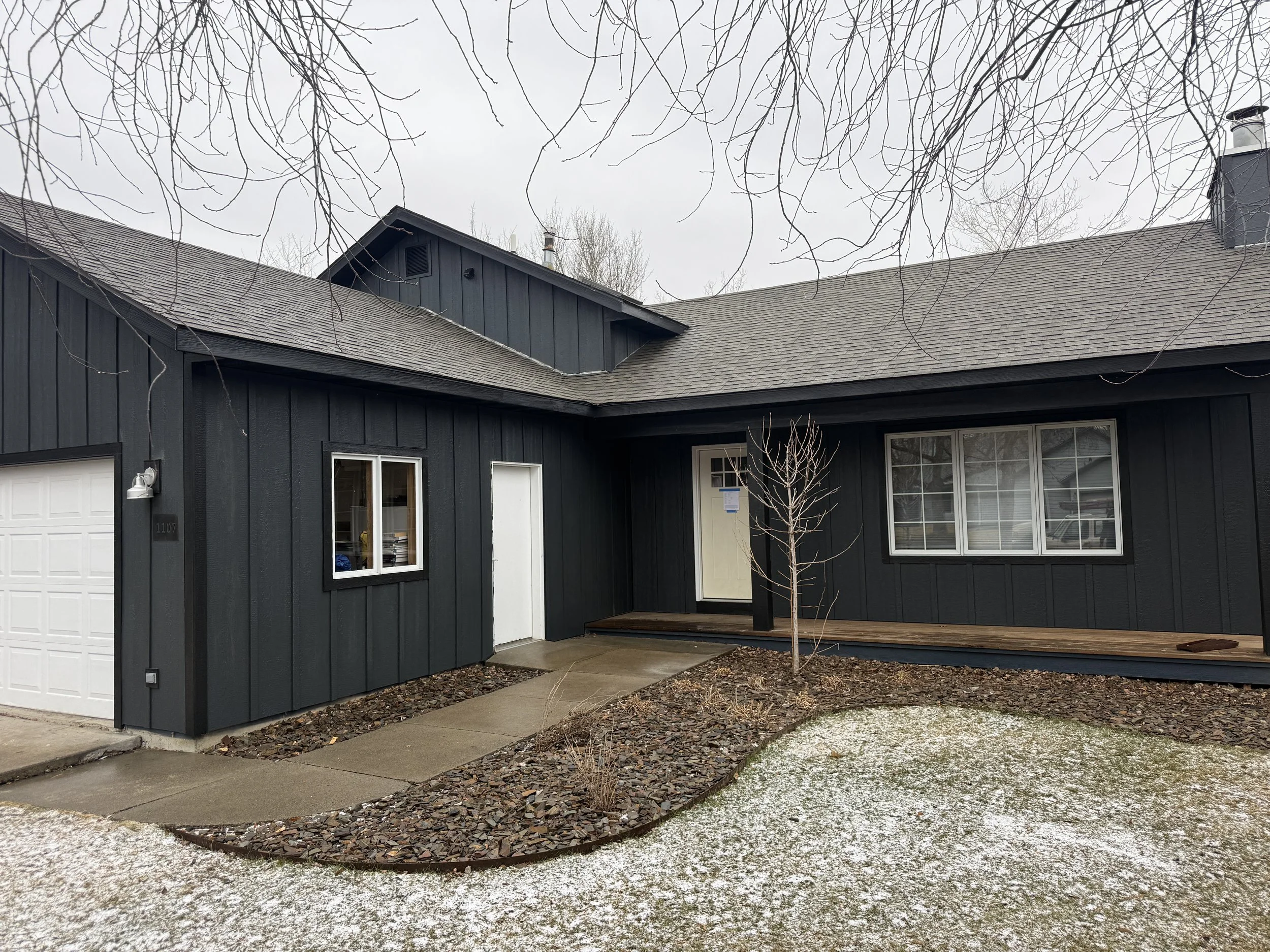 A black house with a white garage door, a small window, a white door, and a large window. There is a small leafless tree in front, with a walkway leading to the house, and a light dusting of snow on the ground.