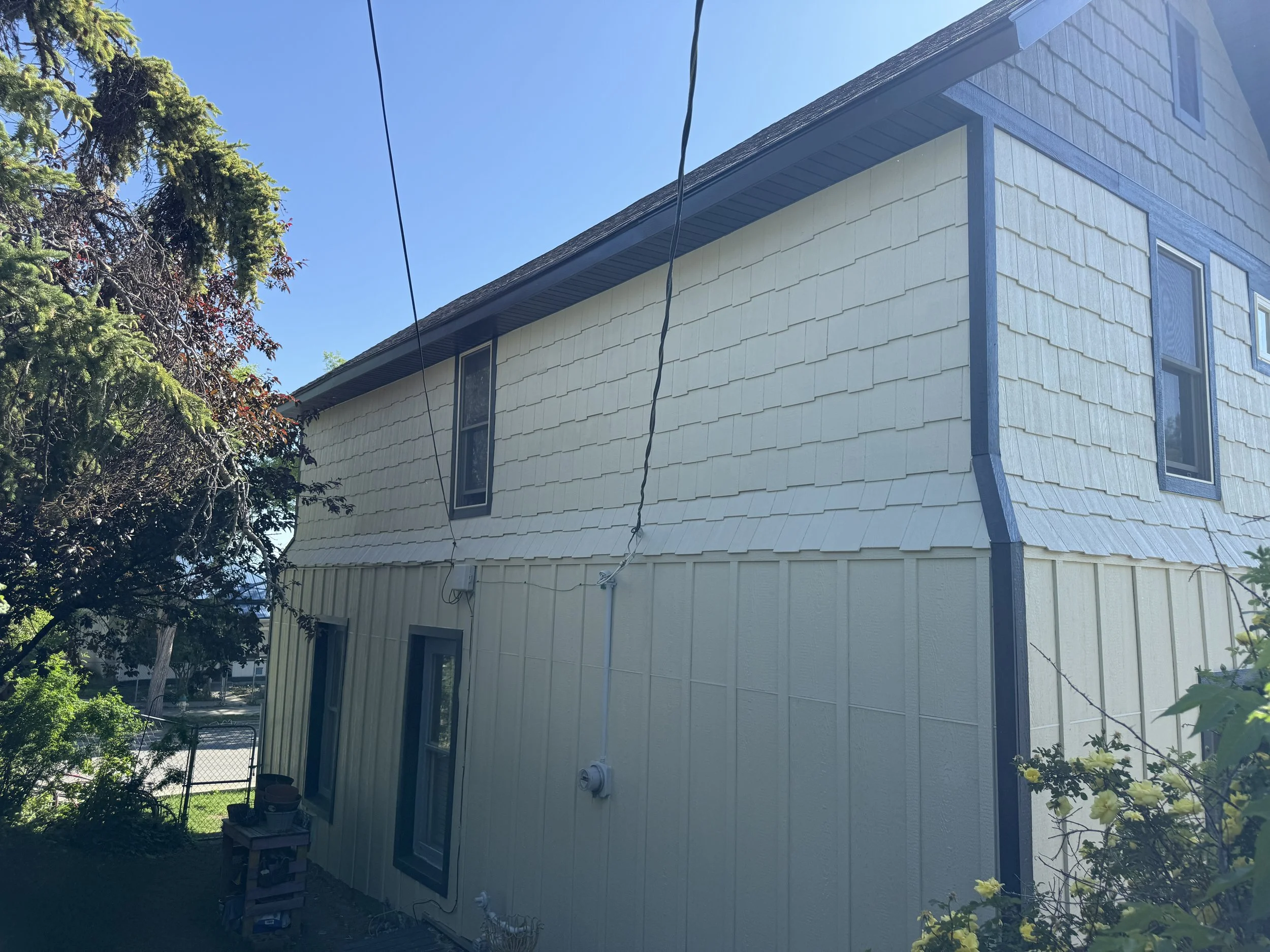 A two-story house with beige siding and dark blue trim, with several windows and a tree on the left side. Clear blue sky in the background.