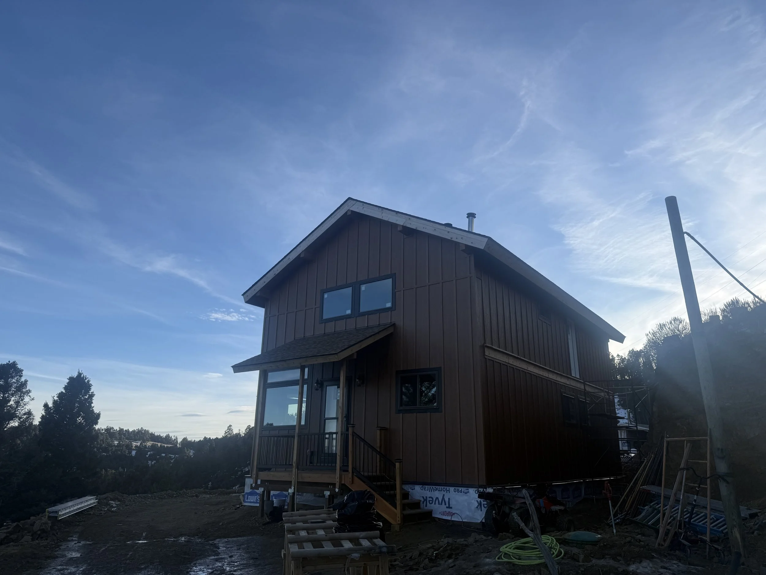 A two-story house under construction with brown siding, a small porch, and a ramp. The sky is partly cloudy.