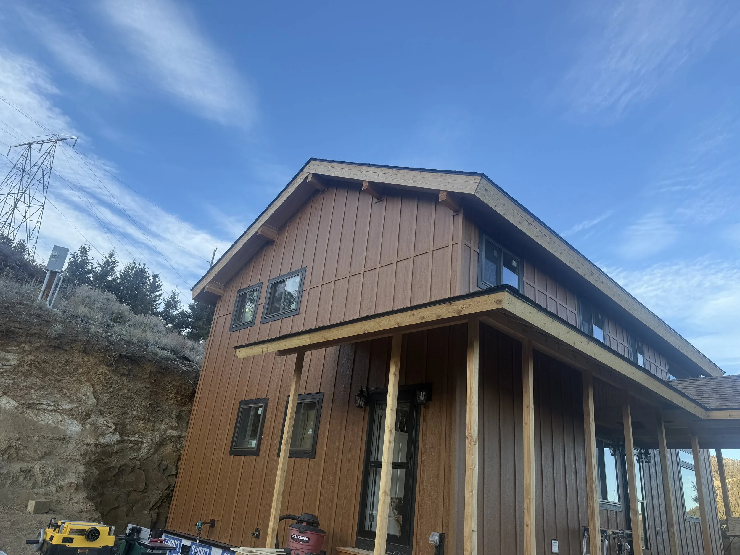 Newly constructed multi-story wooden house with a covered porch, set against a blue sky with wispy clouds and an exposed hillside on the left.