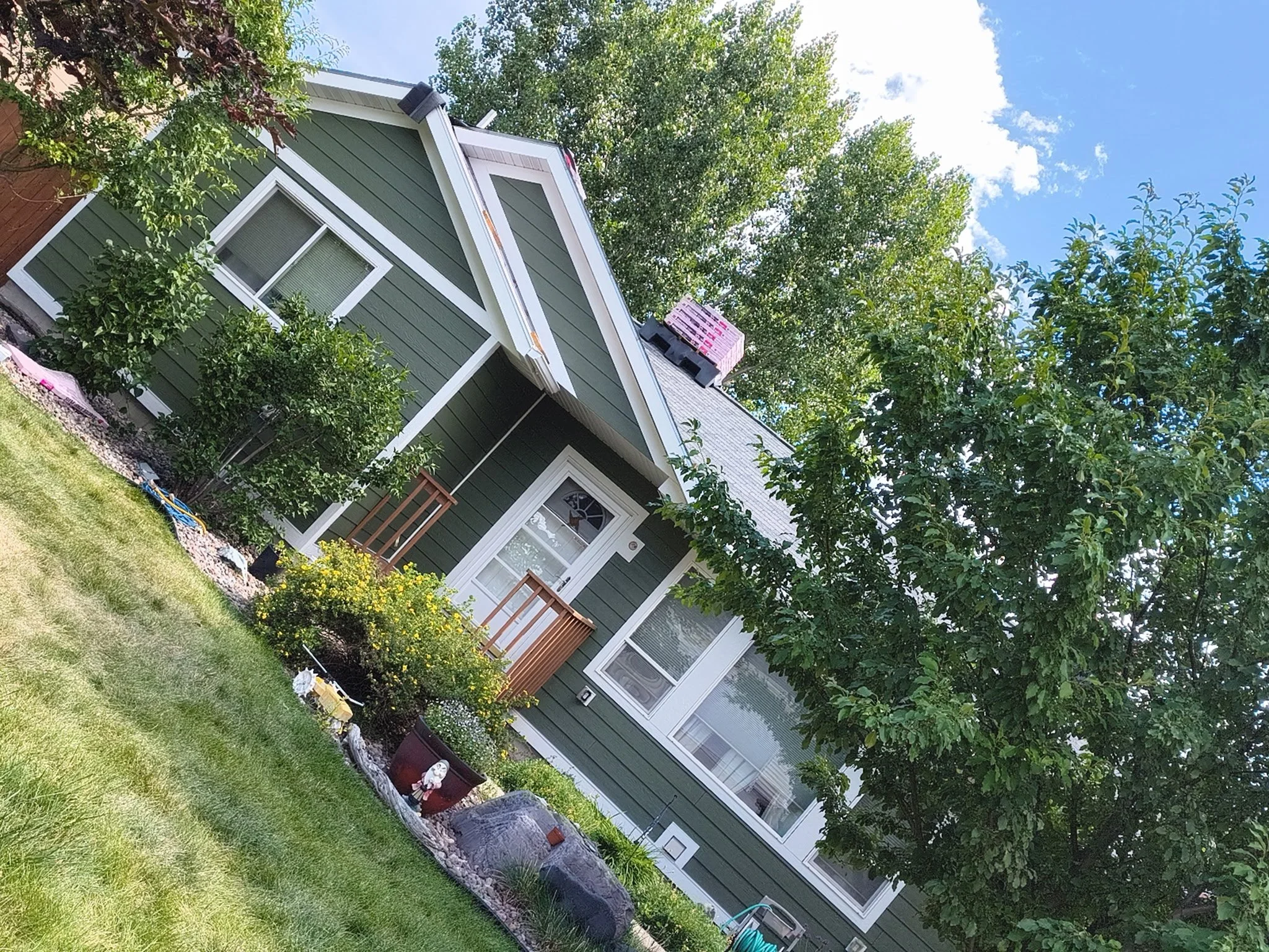 Front yard of a green house with white trim, multiple windows, small porch with brown railings, trees, and a garden with rocks and plants.