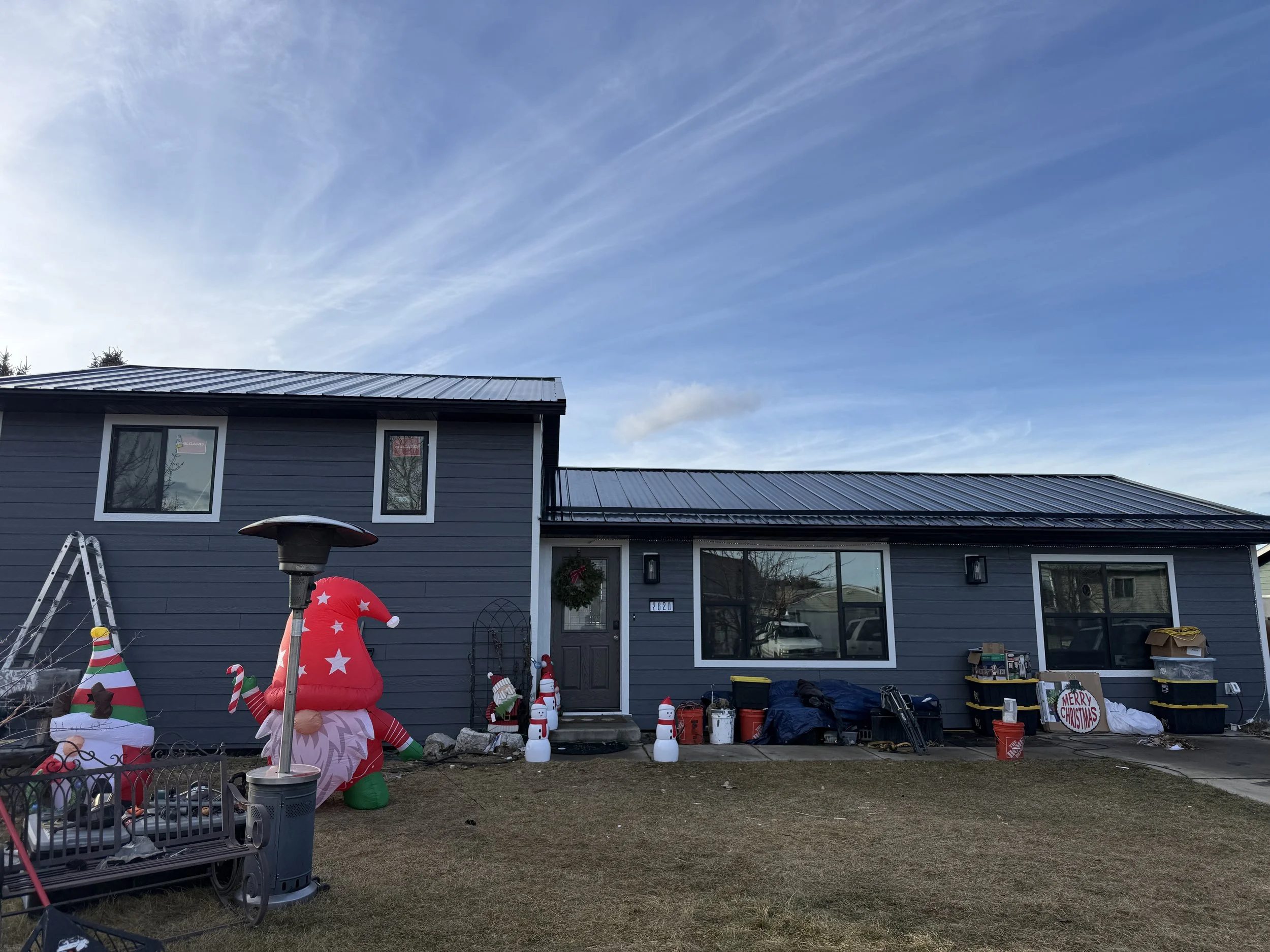 Front yard of a blue house decorated with Christmas inflatables and decorations. There is a large inflatable elf, snowmen, and other holiday decor. The house has a wreath on the door and Christmas signs.