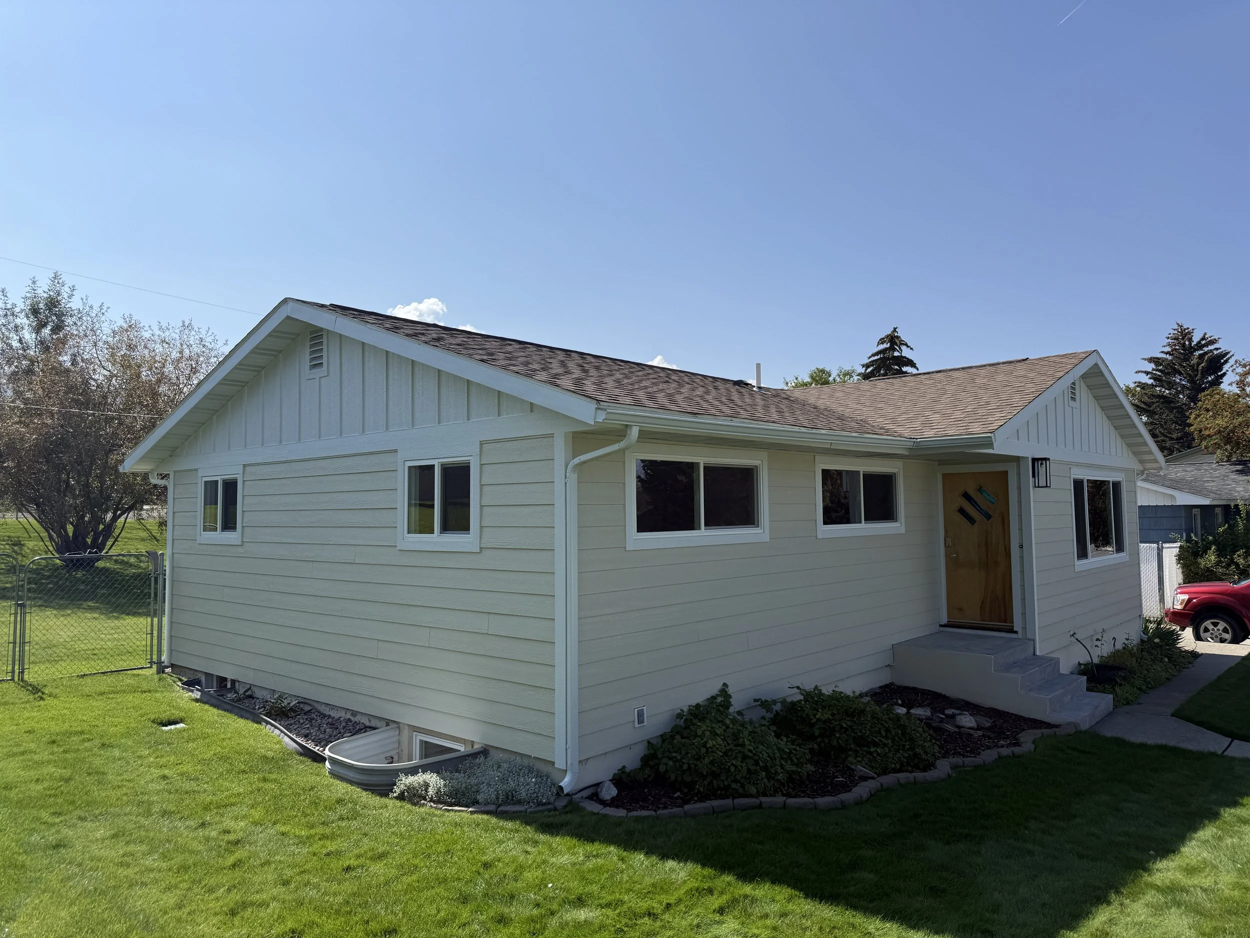 A single-story house with white siding, a gabled roof, and multiple windows. The front door is wooden with diagonal designs, and there are steps leading to the entrance. The house is surrounded by a green lawn and garden plants, with a fence visible 