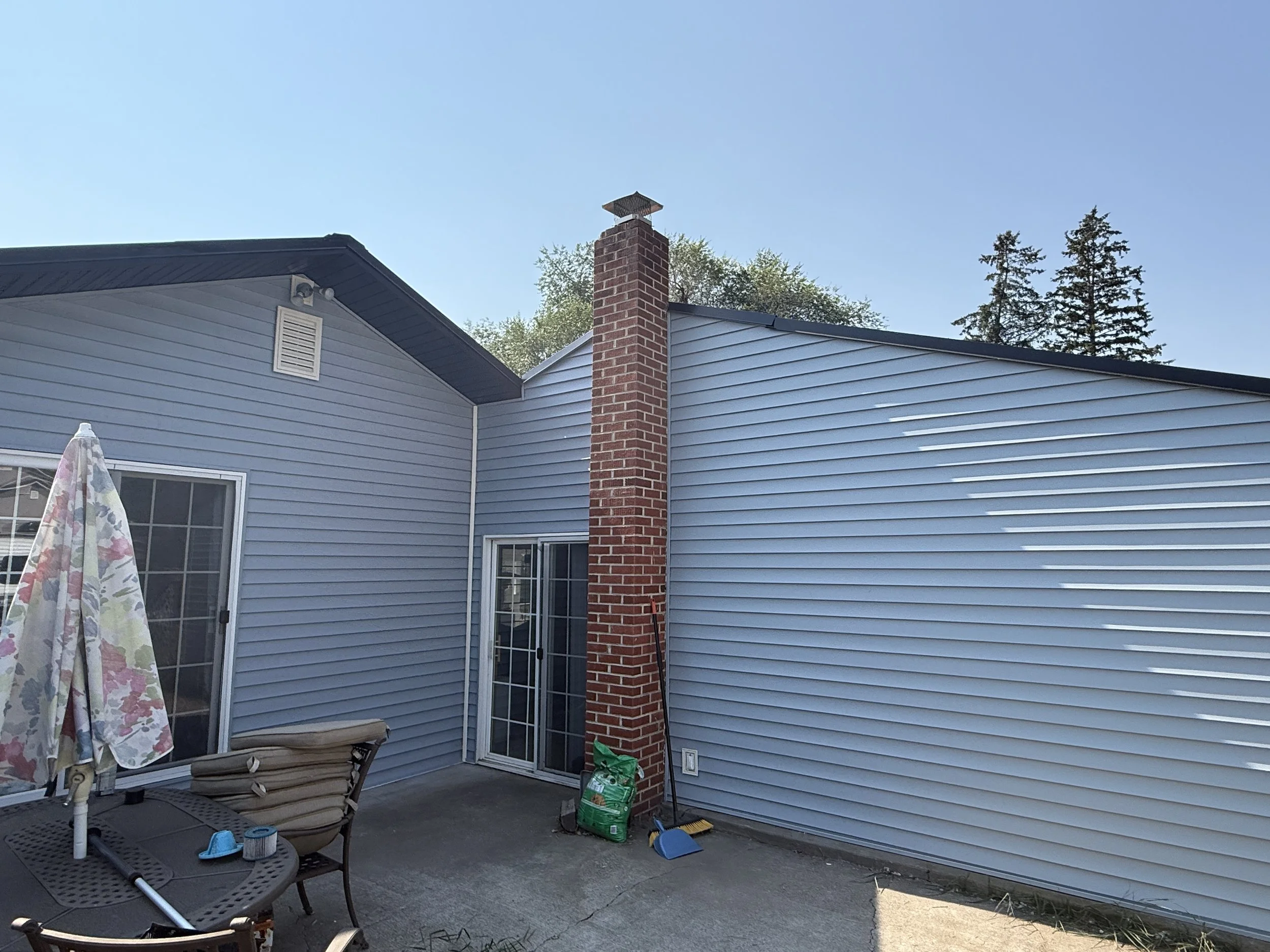 Backyard patio area with a blue house, red brick chimney, patio furniture, and gardening tools on a concrete slab under a clear blue sky.