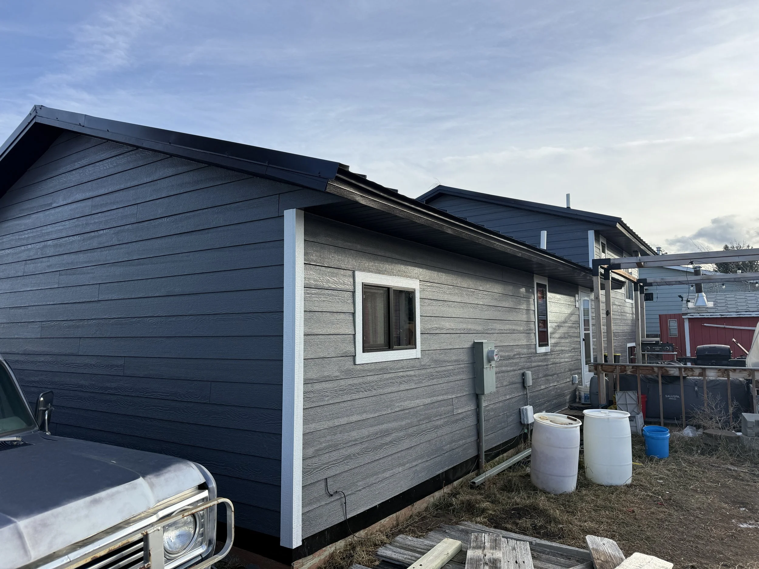 Side view of a gray house with horizontal siding, three small windows, and a sloped roof. In the foreground, a part of a truck is visible, and there are white and blue barrels and various outdoor items around the house.