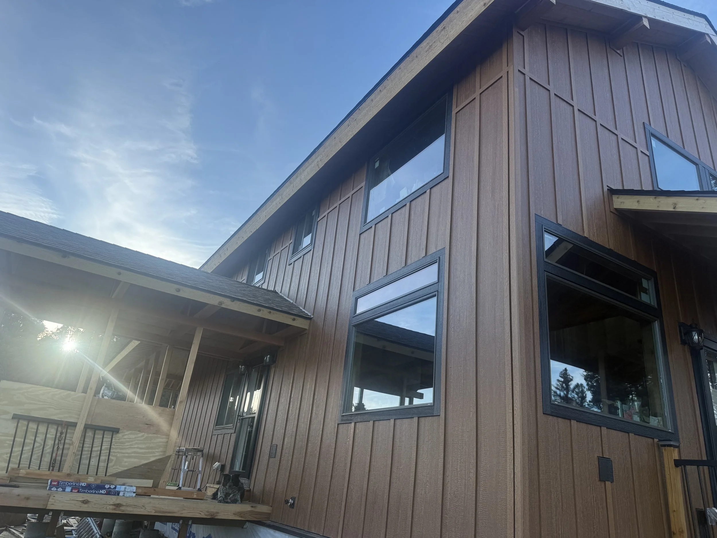 Newly constructed two-story house with wooden exterior siding, multiple large windows, and a covered porch area. The sky is clear with some clouds, and sunlight is shining from the left side of the image.