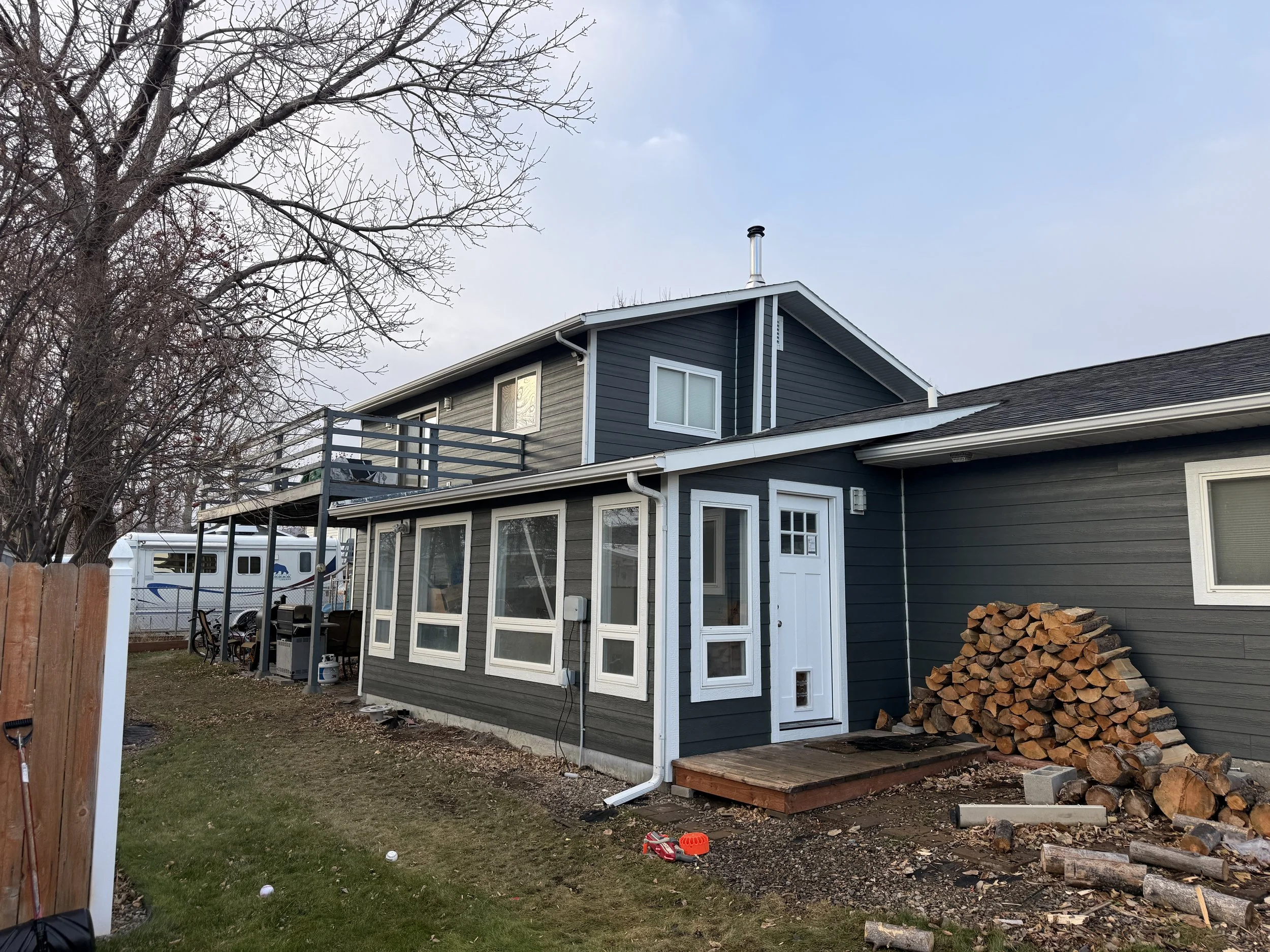 A two-story house with dark gray siding, white trim, and a small deck on the upper level. A pile of firewood is stacked next to the house, and a small wooden platform leads to a door. There are bare trees and a RV in the background.